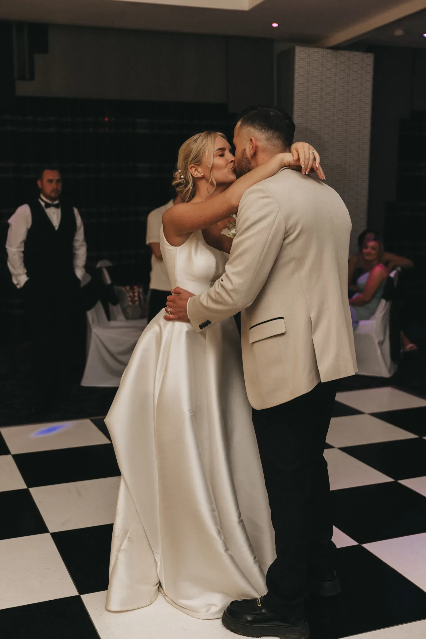 A bride in a white gown kisses a groom in a beige suit on a black and white checkered dance floor, with wedding guests in the background.