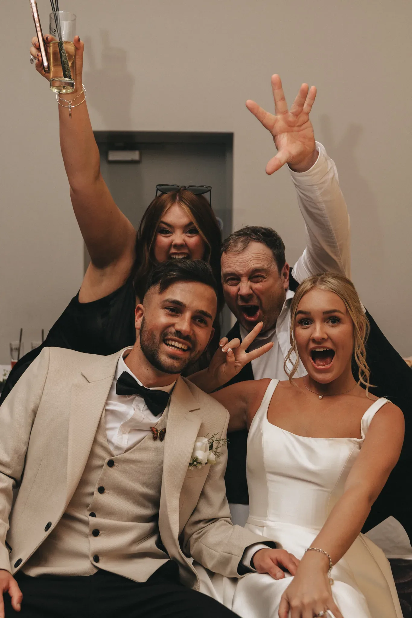 A joyful group of four at a wedding, with a bride and groom in the front and two guests behind them. everyone is smiling and making playful gestures, the groom in a white tuxedo and the bride in a white dress.