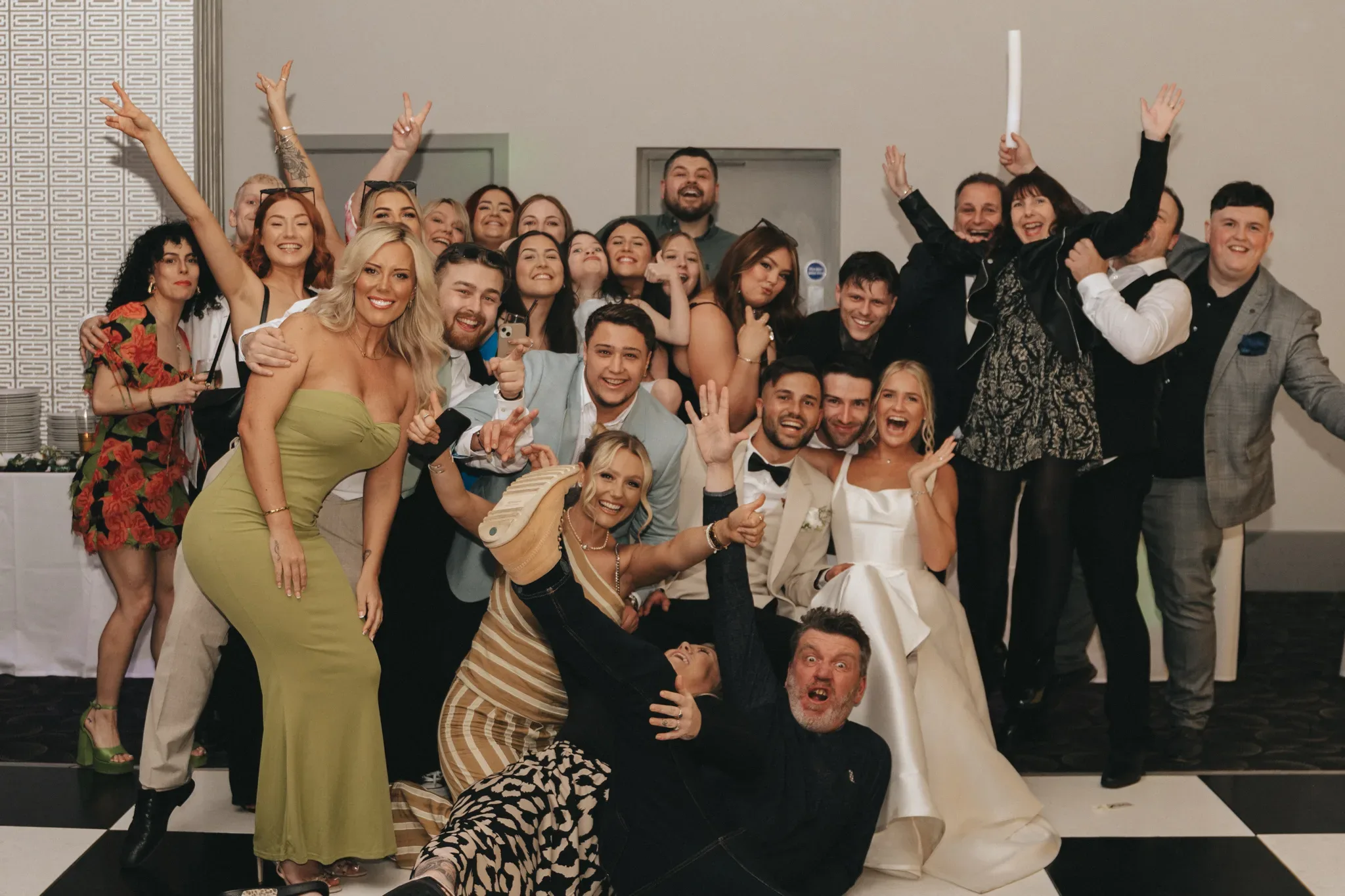 A lively group of around twenty people, including a bride in white and a groom in a suit, celebrates joyfully at a wedding reception. they're posing playfully, some raising their arms or making faces, with a neutral-toned wall in the background.