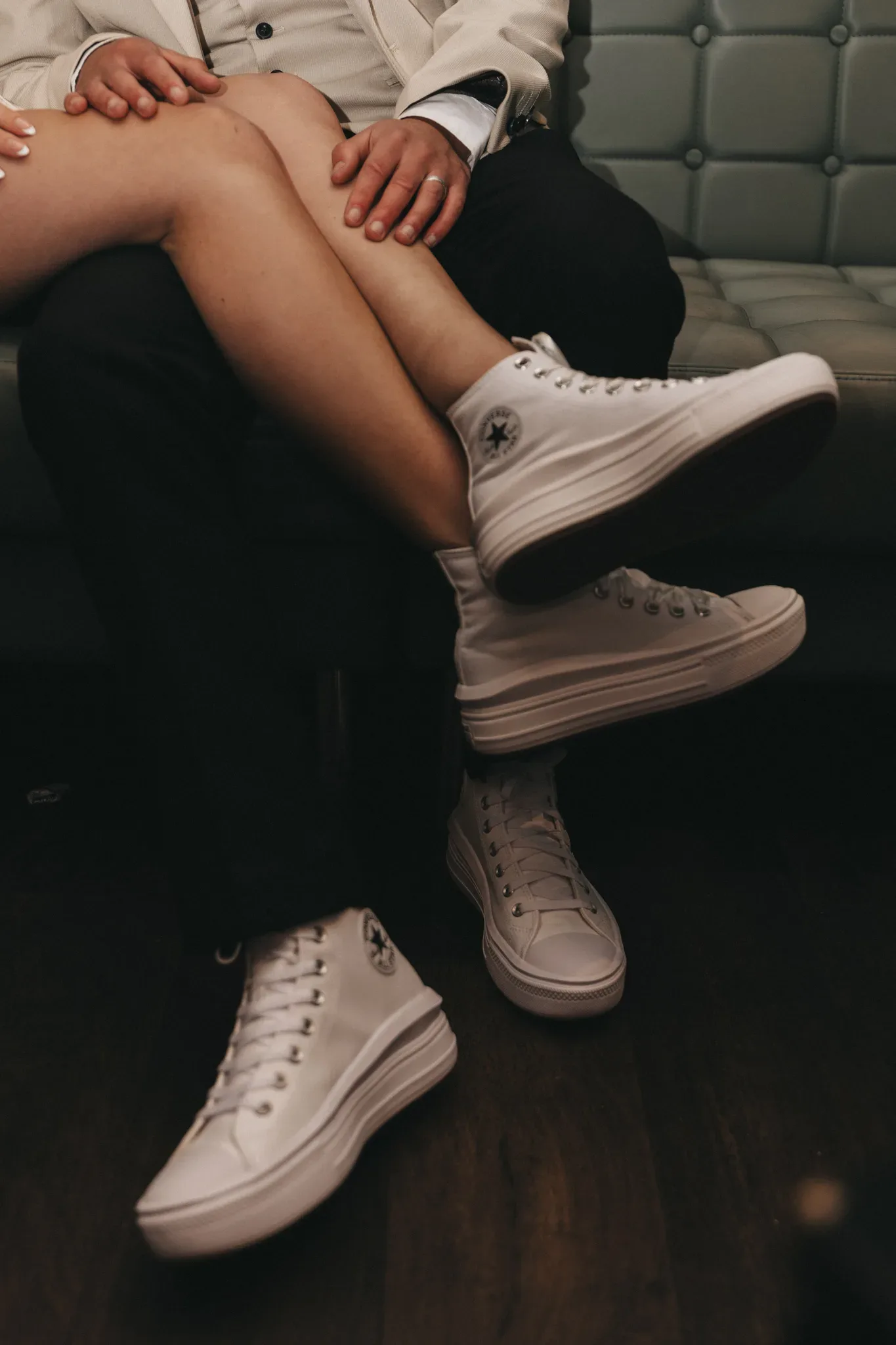 A couple sits closely on a green tufted couch, with focus on their white converse sneakers. one person wears black pants and the other a cream outfit; their hands gently touch.