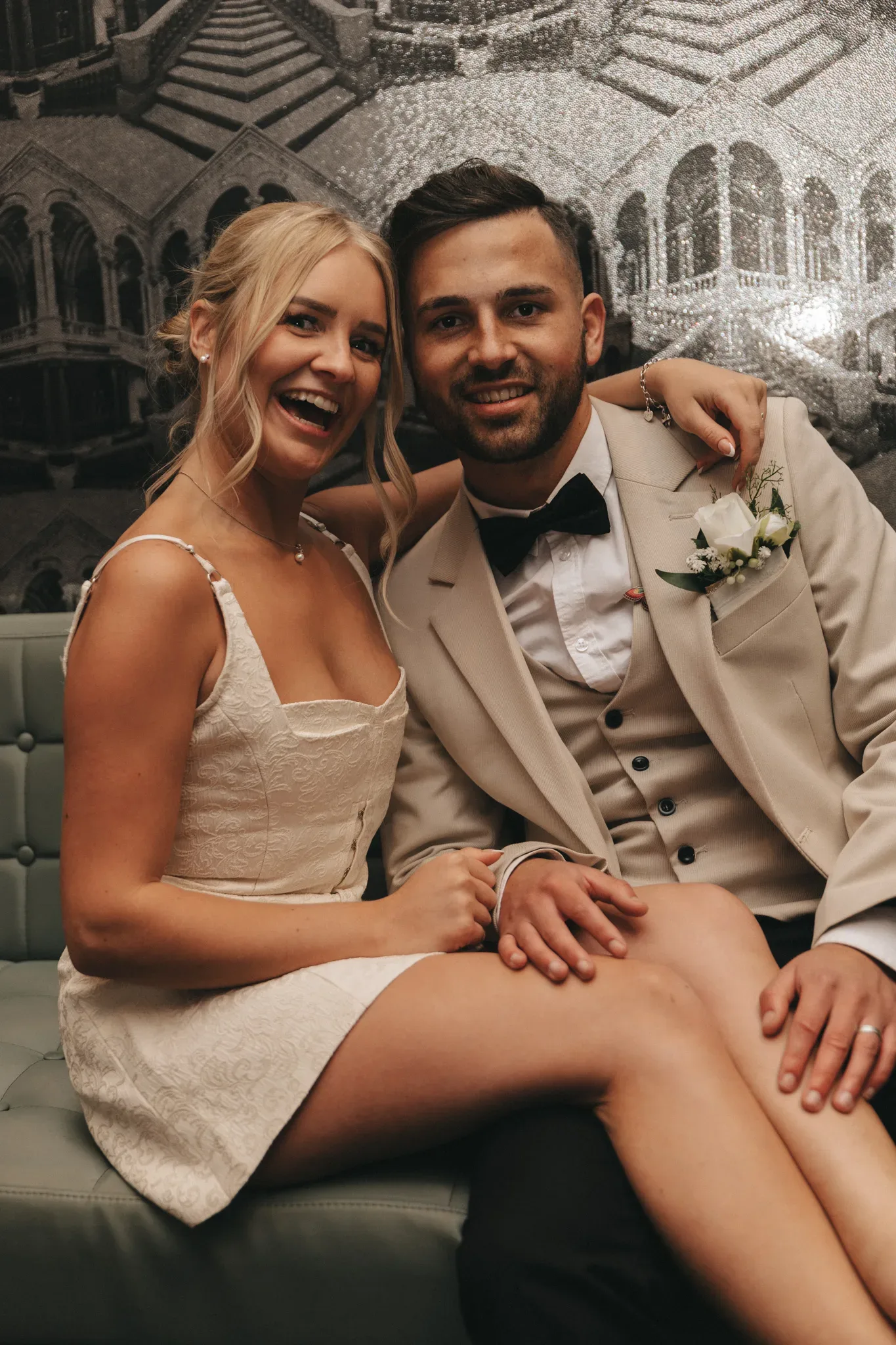 A joyful bride and groom sitting closely on a green velvet sofa. the bride, in a white lace dress, laughs beside the groom, who wears a stylish beige suit with a boutonniere. a glittering chandelier backdrop enhances the festive ambiance.