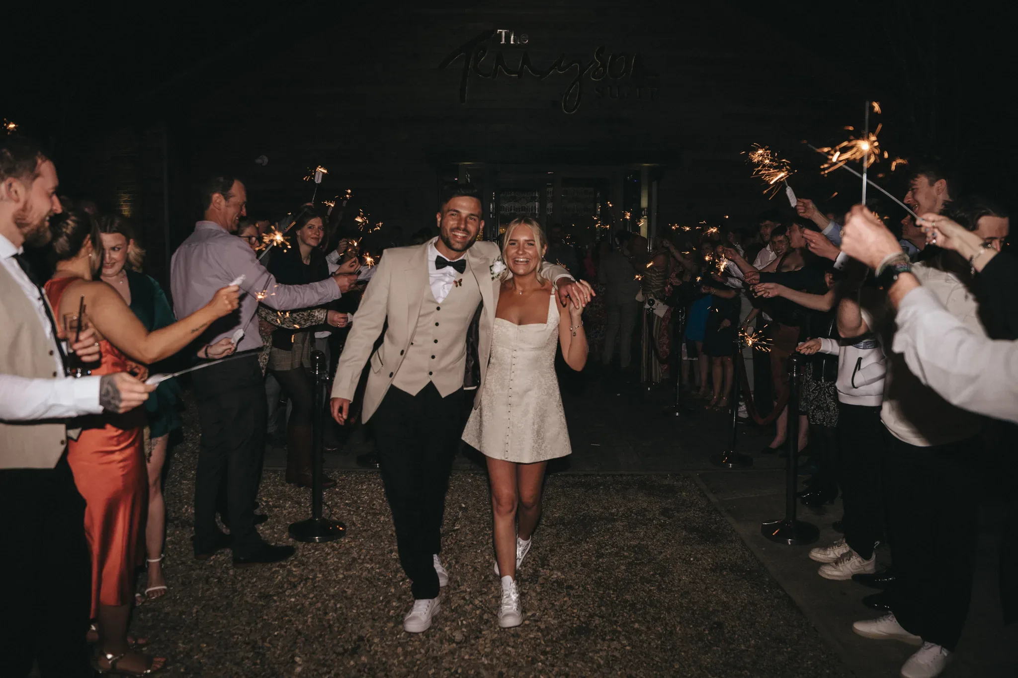A newlywed couple walks through a sparkler send-off at night, smiling joyously. the bride, in a short white dress, and the groom, in a cream suit, hold hands under a dimly lit wooden venue with guests around them holding sparklers.