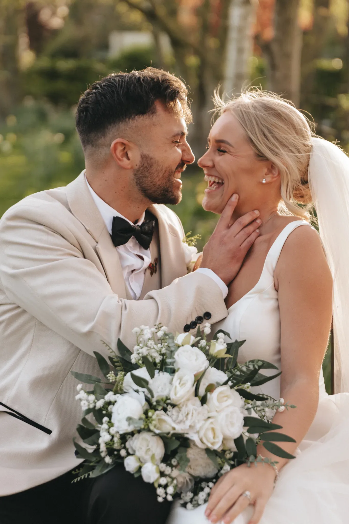 A joyful bride and groom in wedding attire share a close moment, smiling at each other outdoors. the bride holds a bouquet of white flowers. sunlight filters through trees in the background.