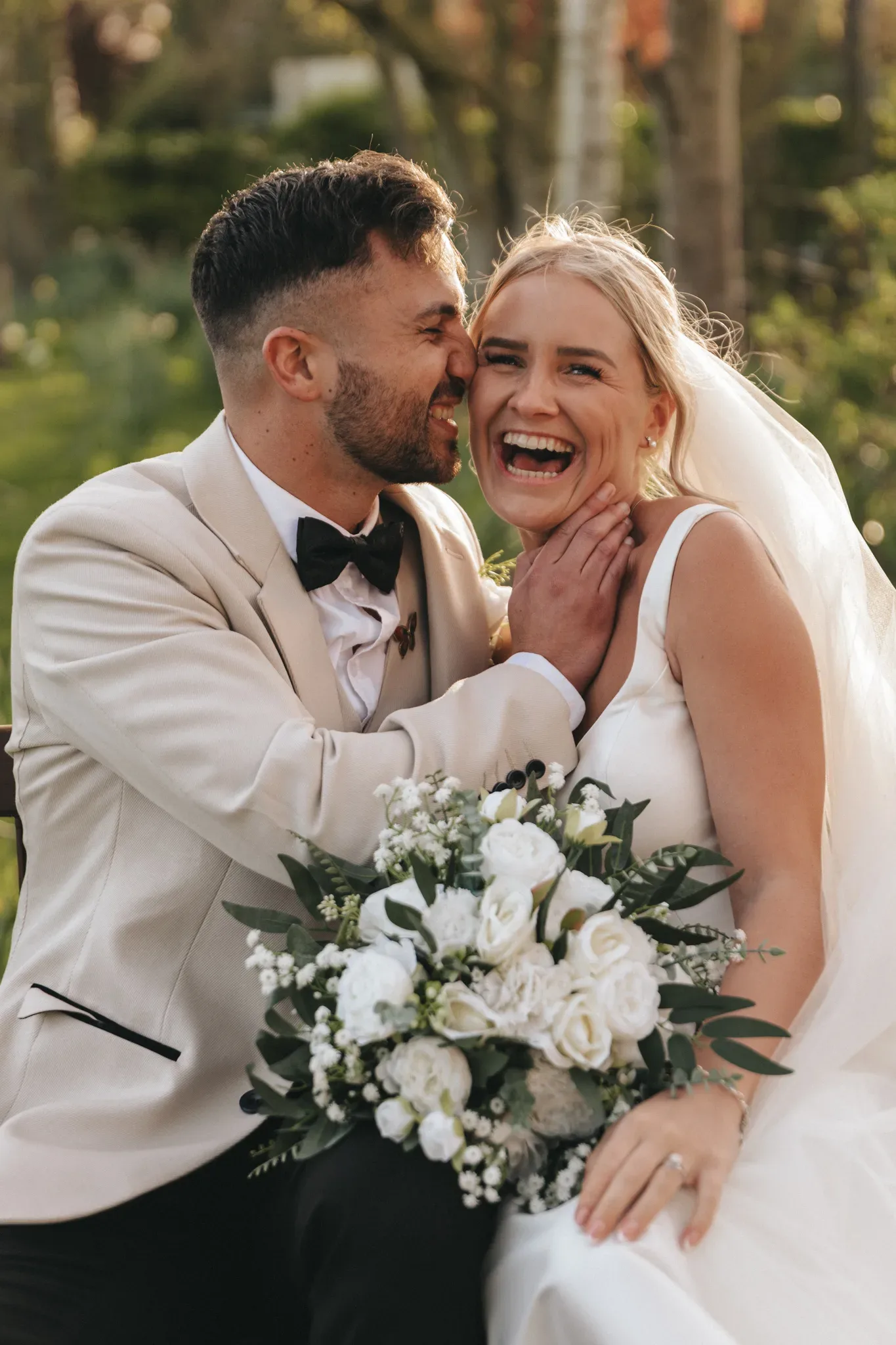 A joyful bride and groom laugh together outdoors. the bride, holding a bouquet of white flowers, wears a white gown, and the groom is in a cream suit and black bow tie. sunlight filters through trees in the background.