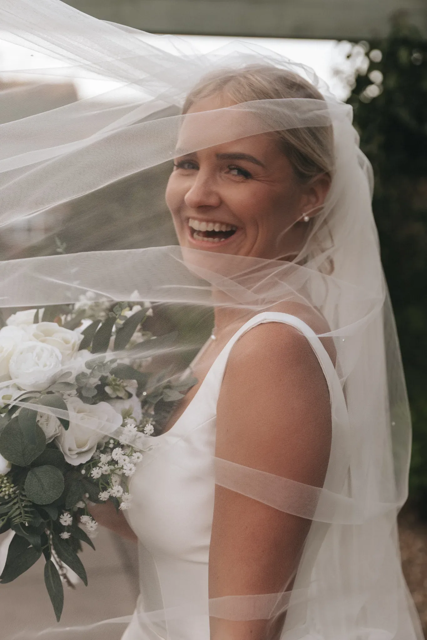 A joyful bride with blonde hair, covered by a translucent veil, smiles broadly, holding a bouquet of white flowers and greenery. she is wearing a white sleeveless dress, with soft, natural light illuminating her.