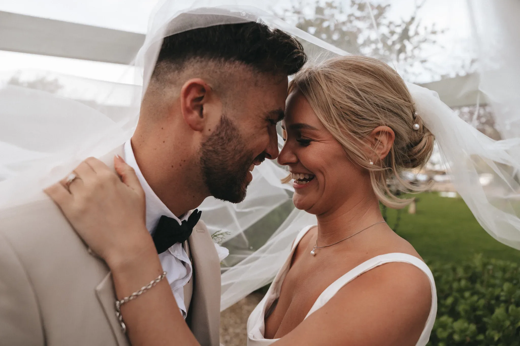 A joyful bride and groom embrace under a sheer veil, their faces close together smiling. the groom is bearded, and the bride has her hair styled up. they are outdoors, with soft, blurred greenery in the background.