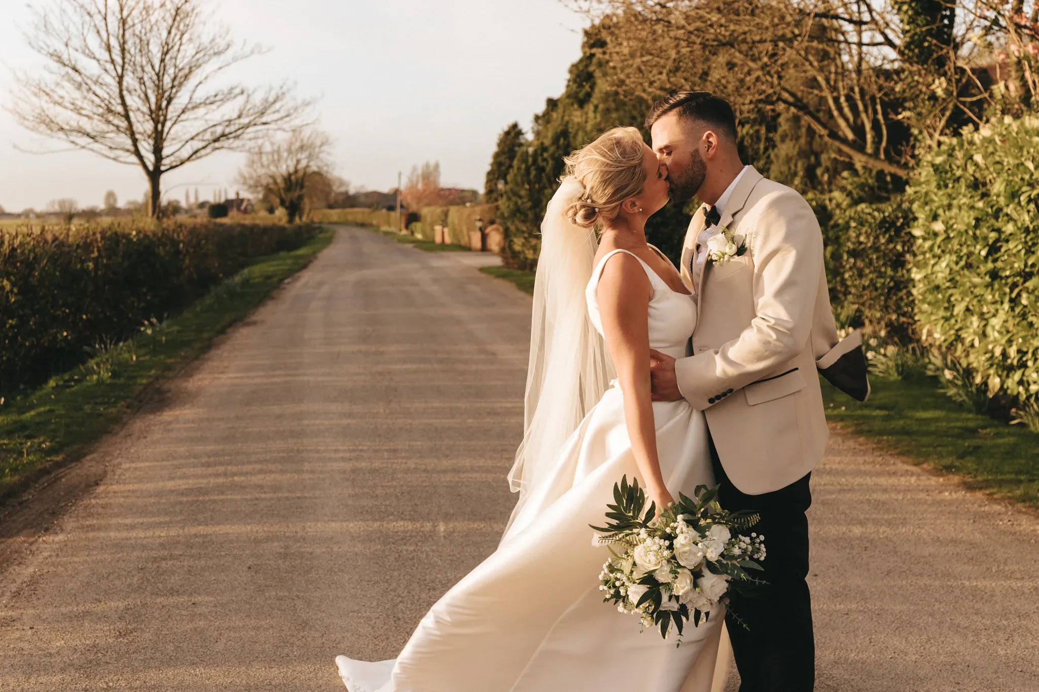 A newlywed couple shares a kiss on a country road. the bride, in a flowing white gown and holding a bouquet, and the groom, in a white suit, are bathed in sunlight with greenery and a clear sky around them.