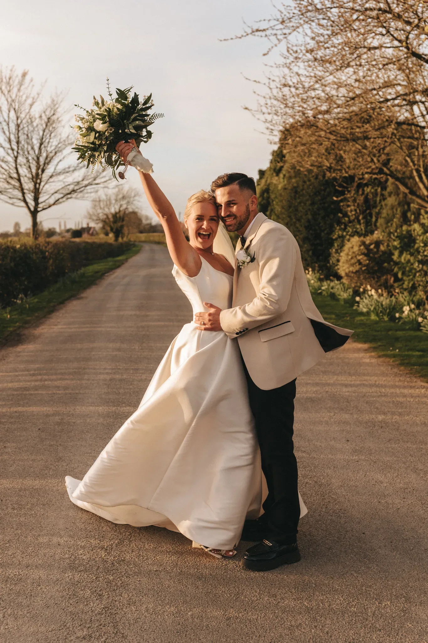 A joyful bride and groom celebrate outdoors on a sunny day. the bride, in a classic white gown, raises a bouquet triumphantly as the groom, in a beige suit, embraces her waist. a rural road with greenery surrounds them.