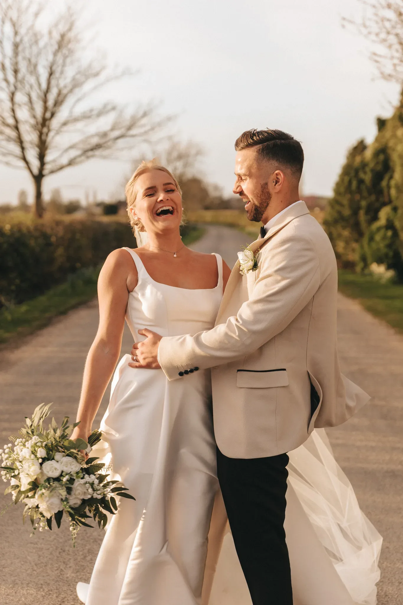 A joyful bride and groom laugh together on a sunlit path. the bride, in an elegant white gown, holds a bouquet; the groom, in a beige suit, gently lifts her. countryside scenery surrounds them.