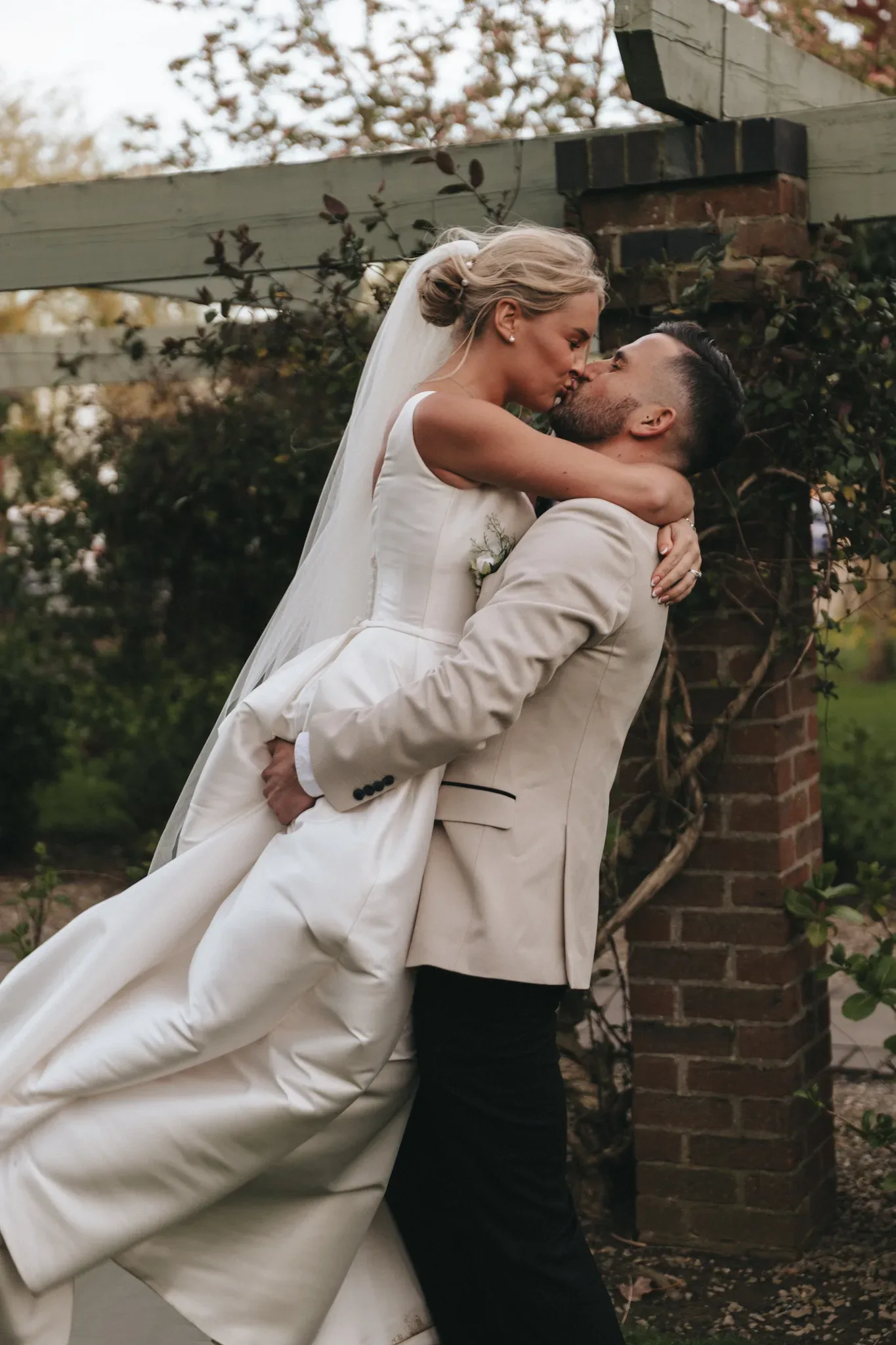 A bride in a white gown and a groom in a beige suit share a passionate kiss. the groom lifts the bride slightly off the ground. they stand under a garden archway surrounded by lush greenery.