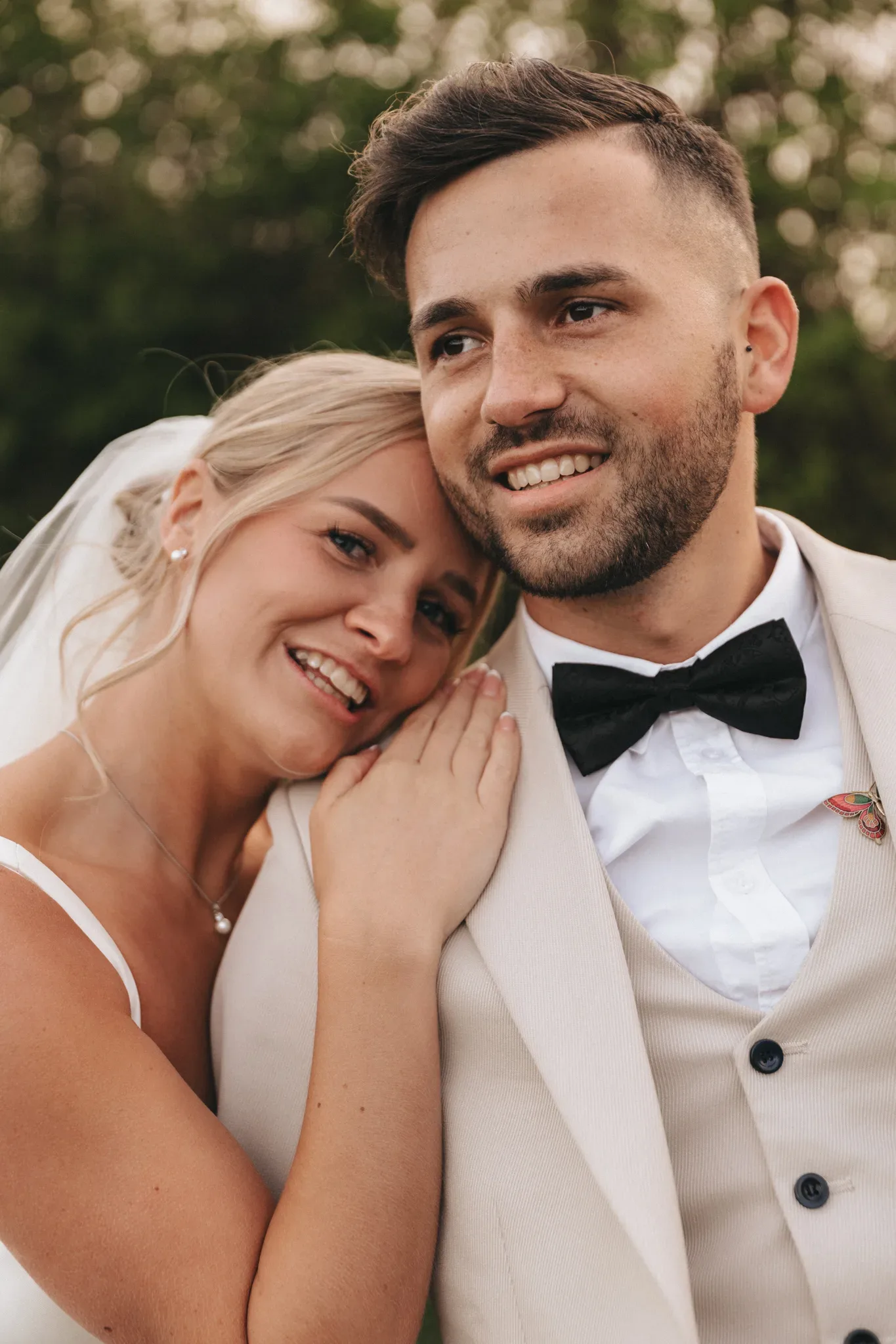 A close-up of a joyful bride and groom. the bride, leaning on the groom's shoulder, is smiling broadly, displaying her blonde hair and white dress. the groom, in a white suit and black bow tie, is glancing away with a subtle smile.