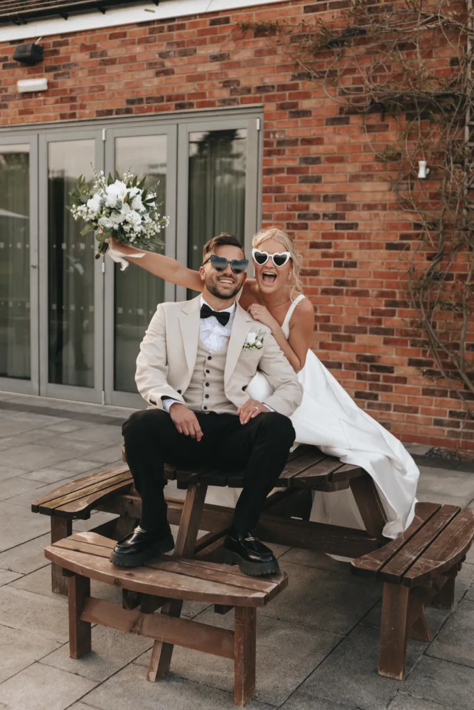 A joyful newlywed couple, a man in a white tuxedo and a woman in a strapless white gown, sitting on a wooden picnic bench, playfully wearing sunglasses and raising a bouquet, with a brick building in the background.