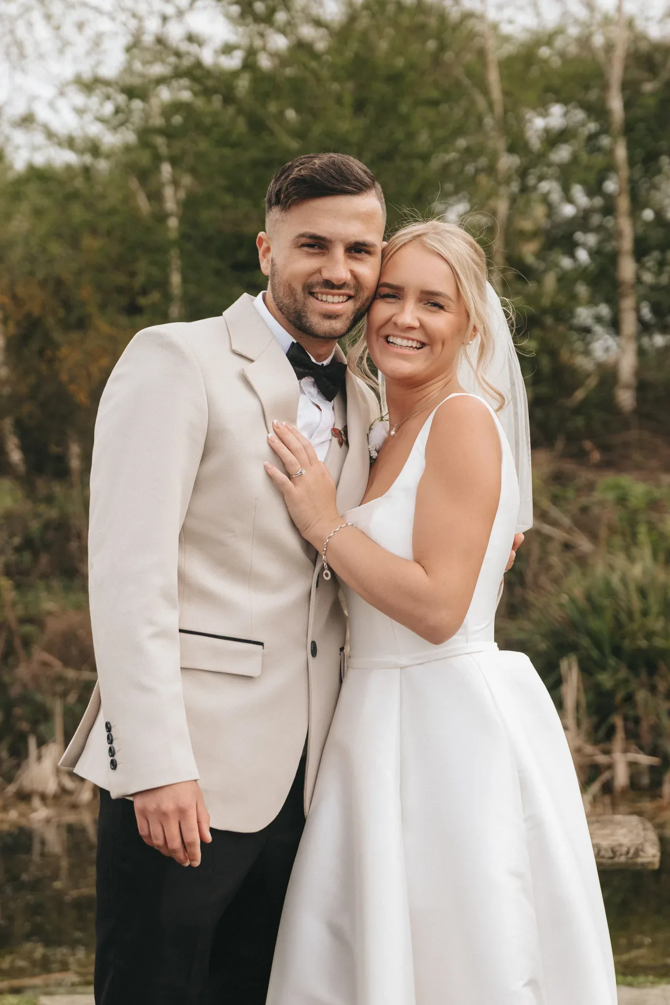 A newlywed couple posing happily outdoors. the groom, in a cream suit, embraces the bride, who is in a classic white dress with a veil. a serene, natural backdrop with soft focus adds to the romantic setting.