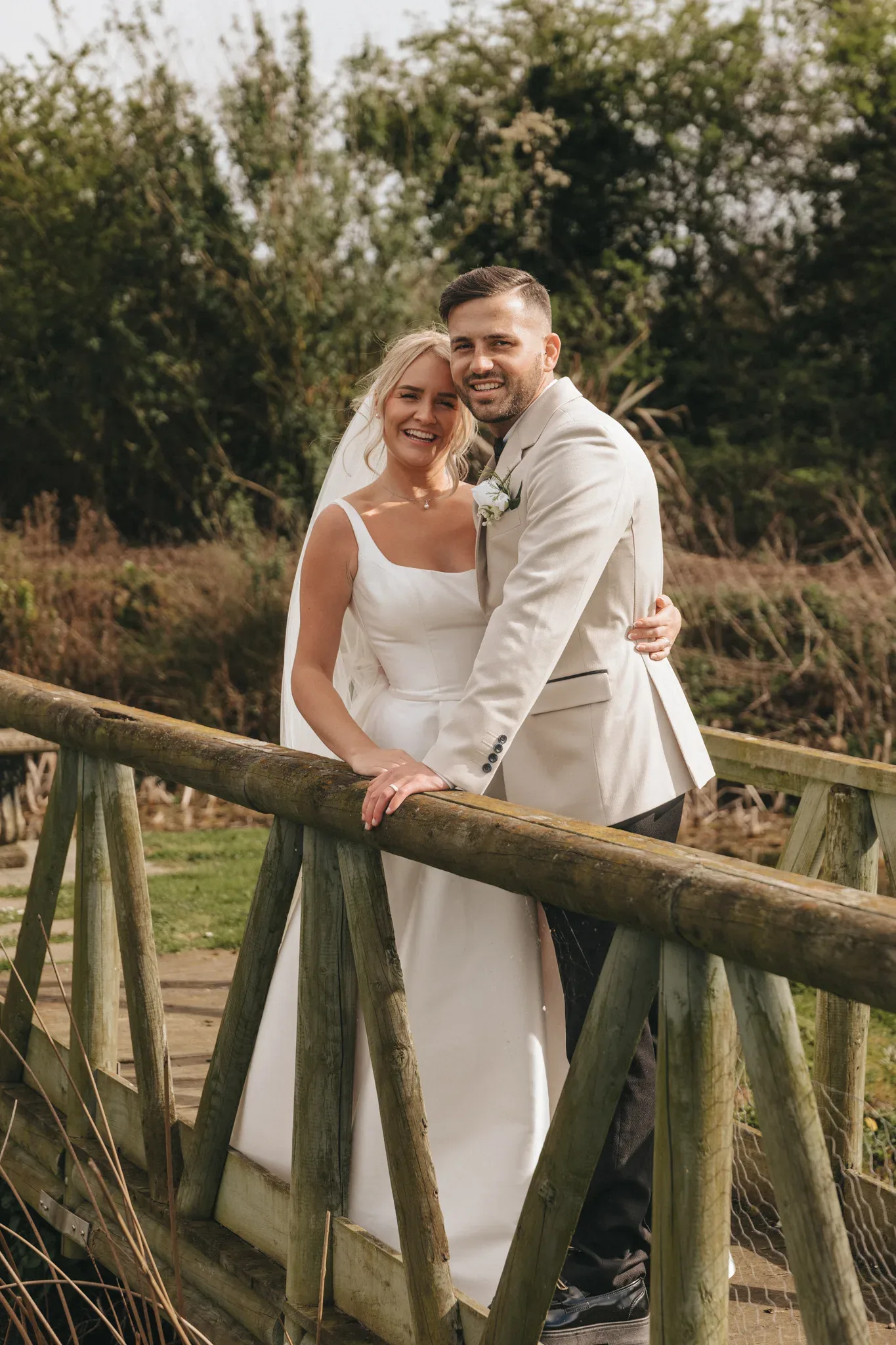 A joyful bride and groom embracing on a wooden bridge in a natural setting, smiling broadly. the bride is in a classic white gown and the groom is in a stylish cream suit.