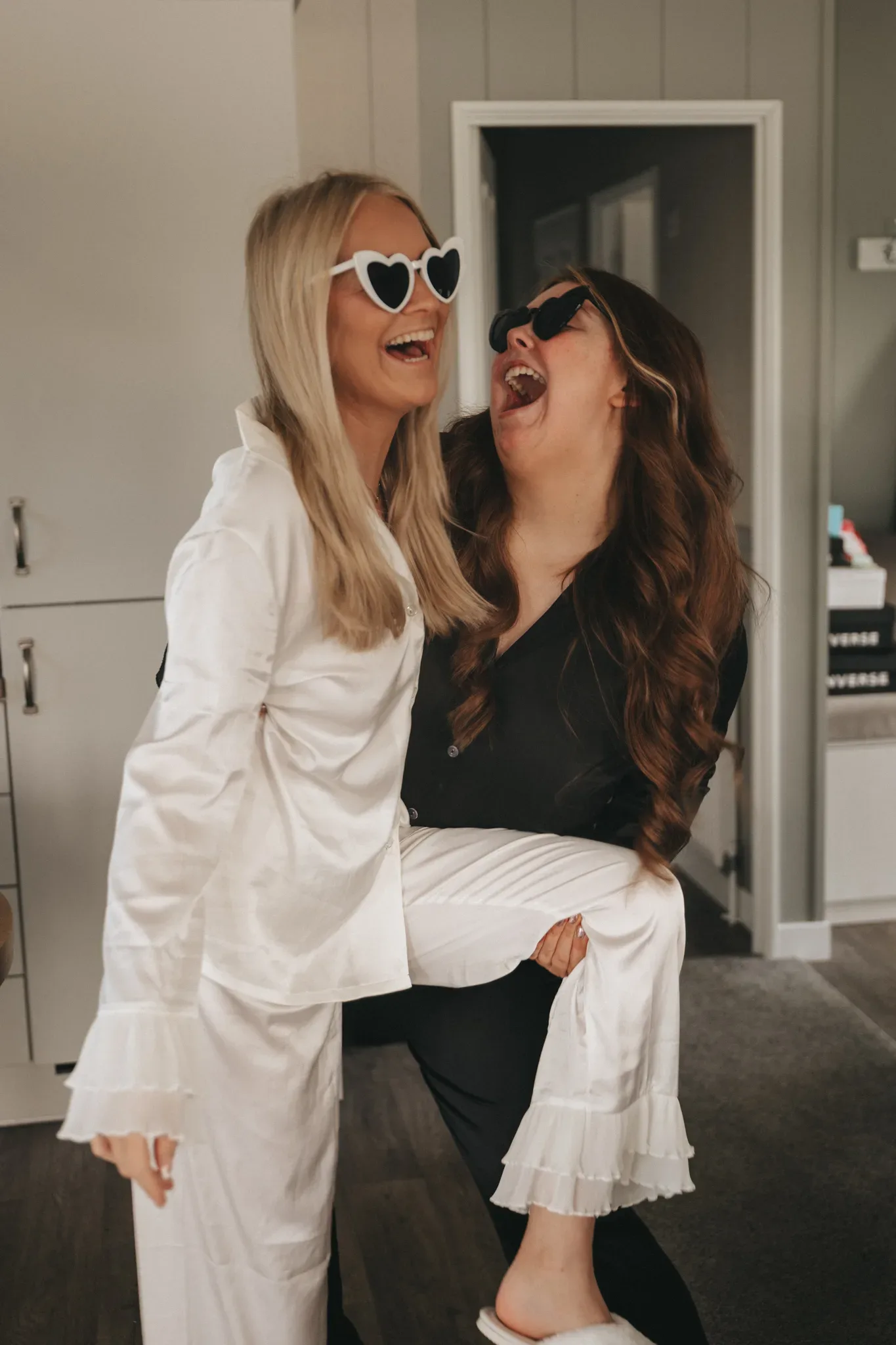 Two women laughing joyfully, one in a white suit lifting the other, who wears a black top and jeans. the woman being lifted wears heart-shaped sunglasses. they are indoors with a modern decor.