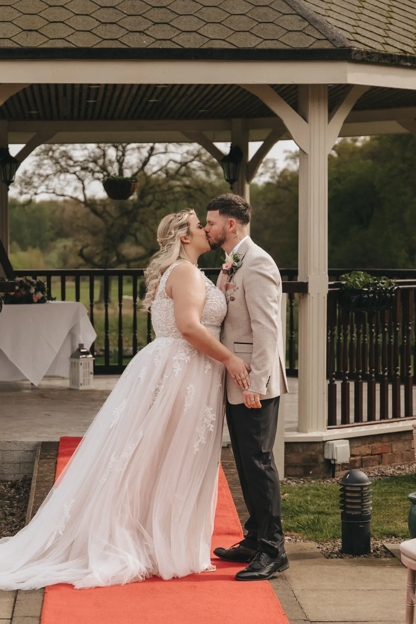 A bride and groom share a kiss under a gazebo. the bride is in a flowing white gown with lace details, and the groom wears a light gray suit with a floral boutonniere. a red carpet and greenery frame the scene.