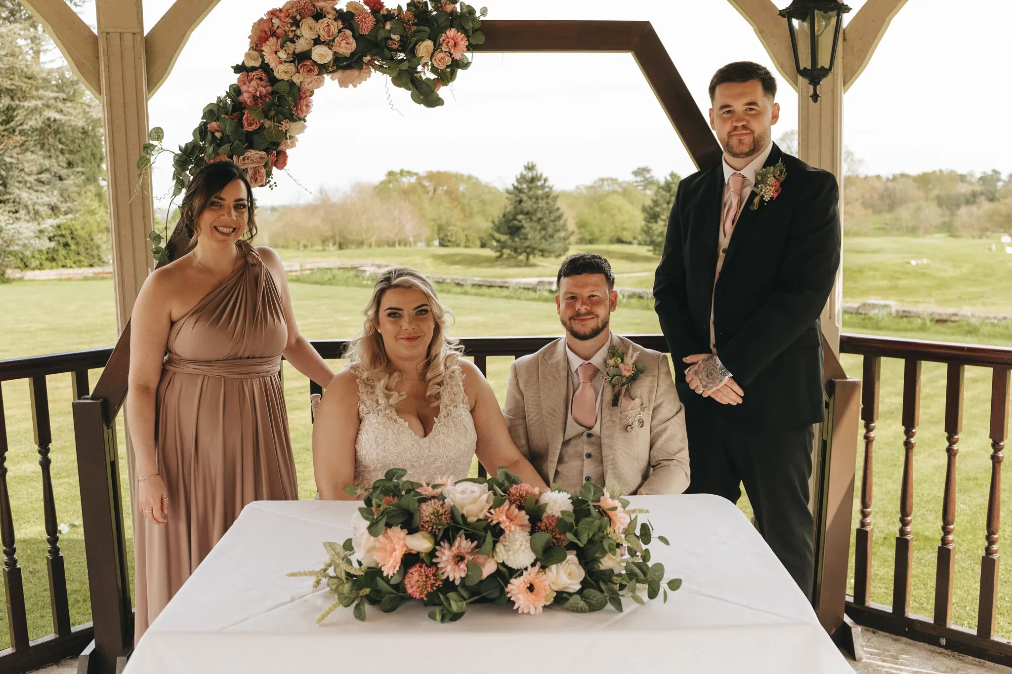 Four people at a wedding ceremony under a gazebo with floral decorations, smiling. two bridesmaids in dresses flank a bride and groom seated at a table adorned with a floral centerpiece.