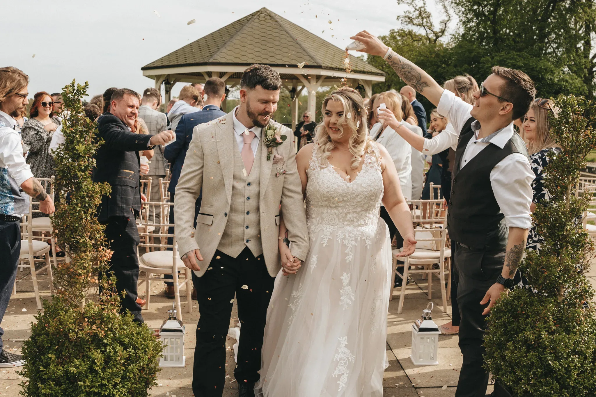 A newlywed couple walks down the aisle outdoors, smiling as guests throw flower petals at them. the bride wears a white lace gown and the groom, a beige suit. a gazebo and greenery are in the background.