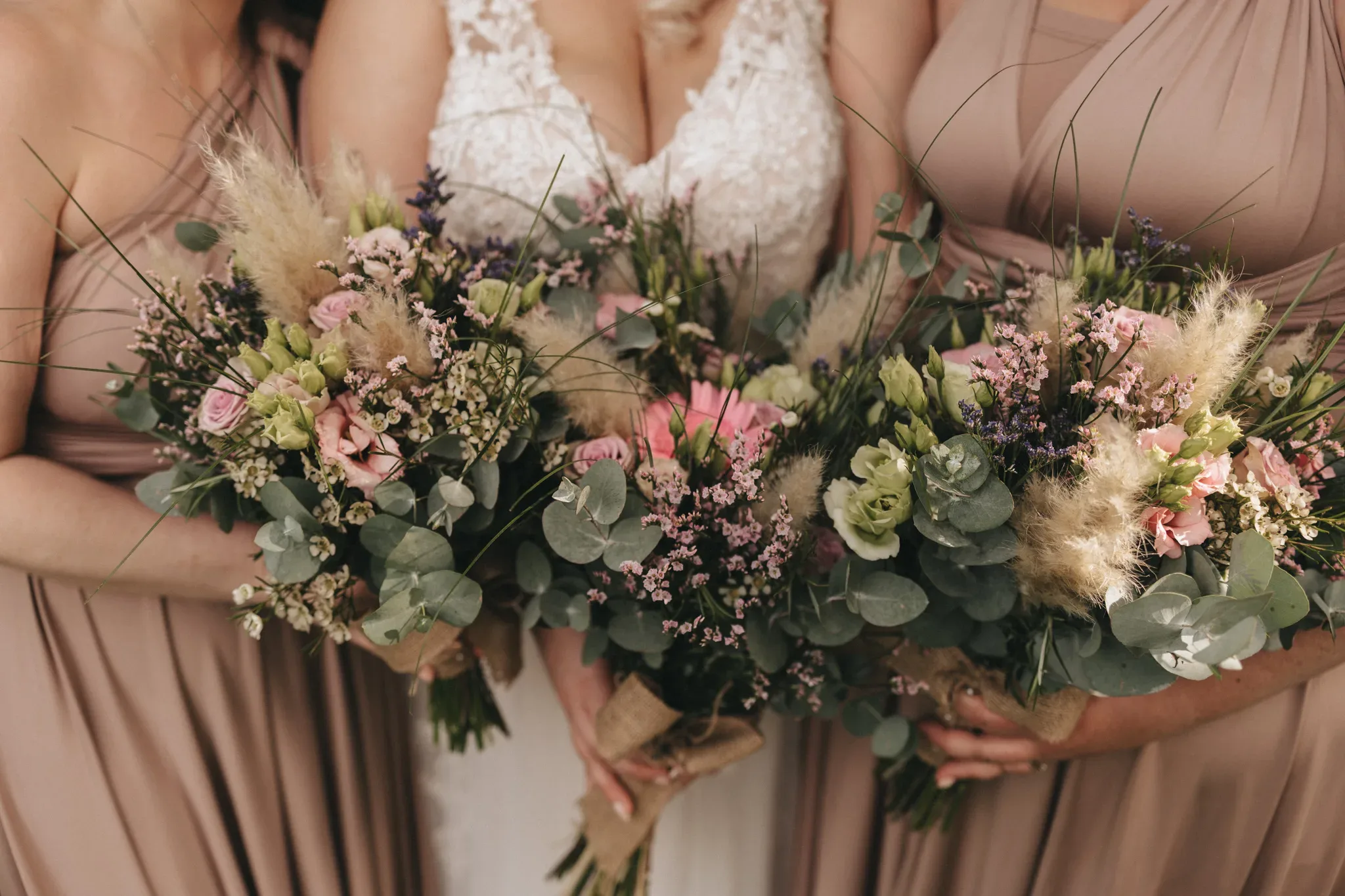 Three women in elegant dresses holding large, rustic bouquets with a mix of pink flowers and greenery, focusing on the bouquets in front of their light-colored dresses.