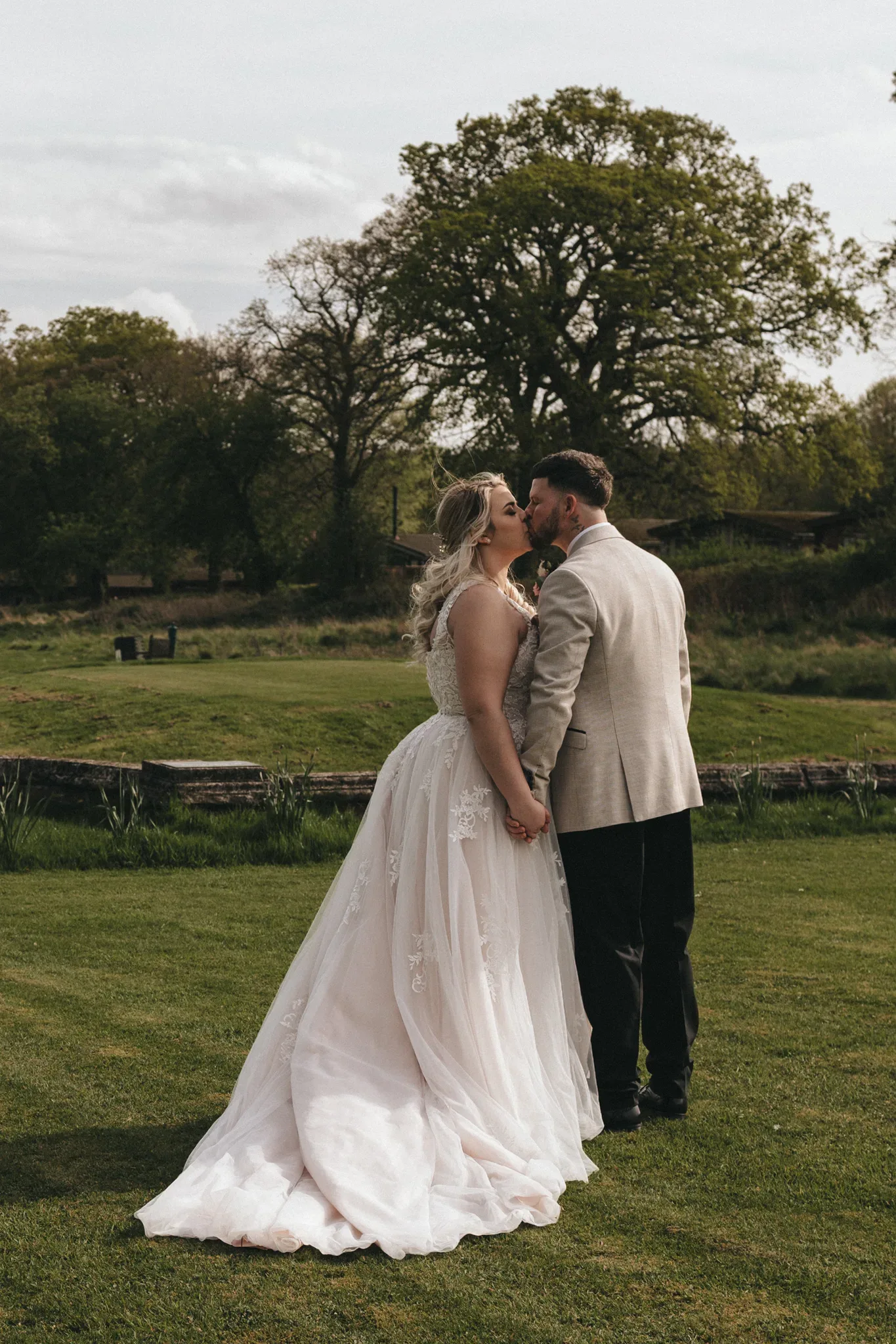 A bride and groom share a kiss in a lush, green meadow. the bride wears a flowing white dress with lace detail, and the groom is in a beige suit. large trees and a clear sky form the backdrop.