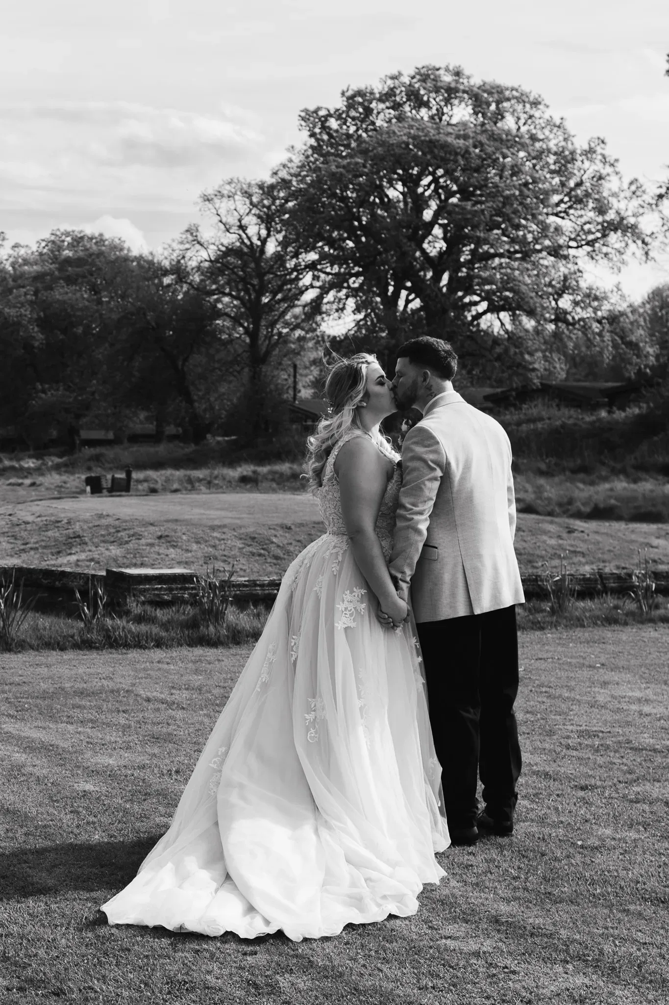 A black and white image of a bride and groom sharing a kiss outdoors. the bride, in a long flowing gown, stands facing away, with the groom in a light suit leaning in for the kiss. serene natural background with a leafy tree and pasture.