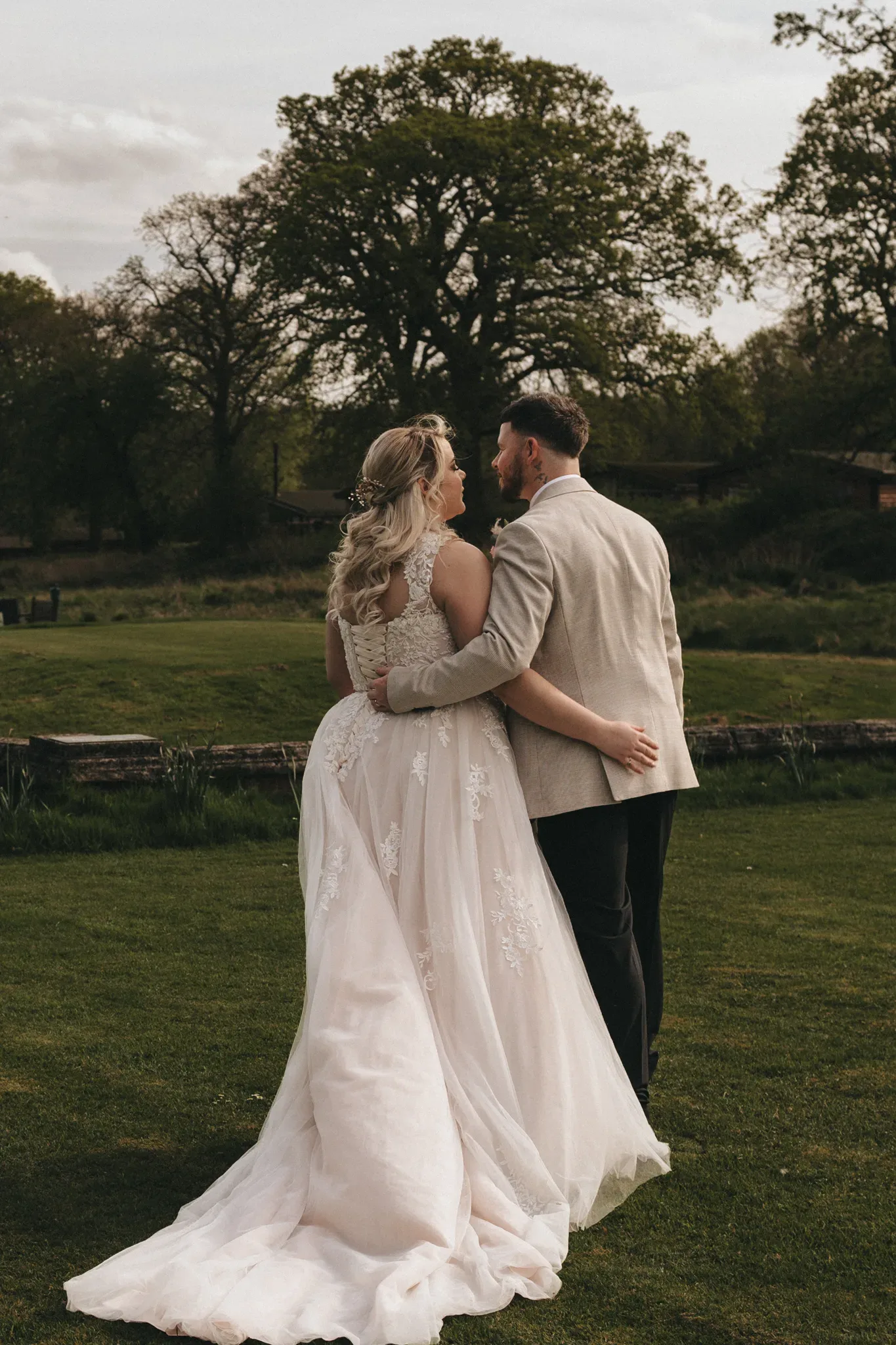A bride and groom embrace outdoors, viewing a natural landscape. the bride's detailed white gown trails behind her, and the groom wears a light-colored suit. they stand on grass near a rustic fence, intimate and serene.