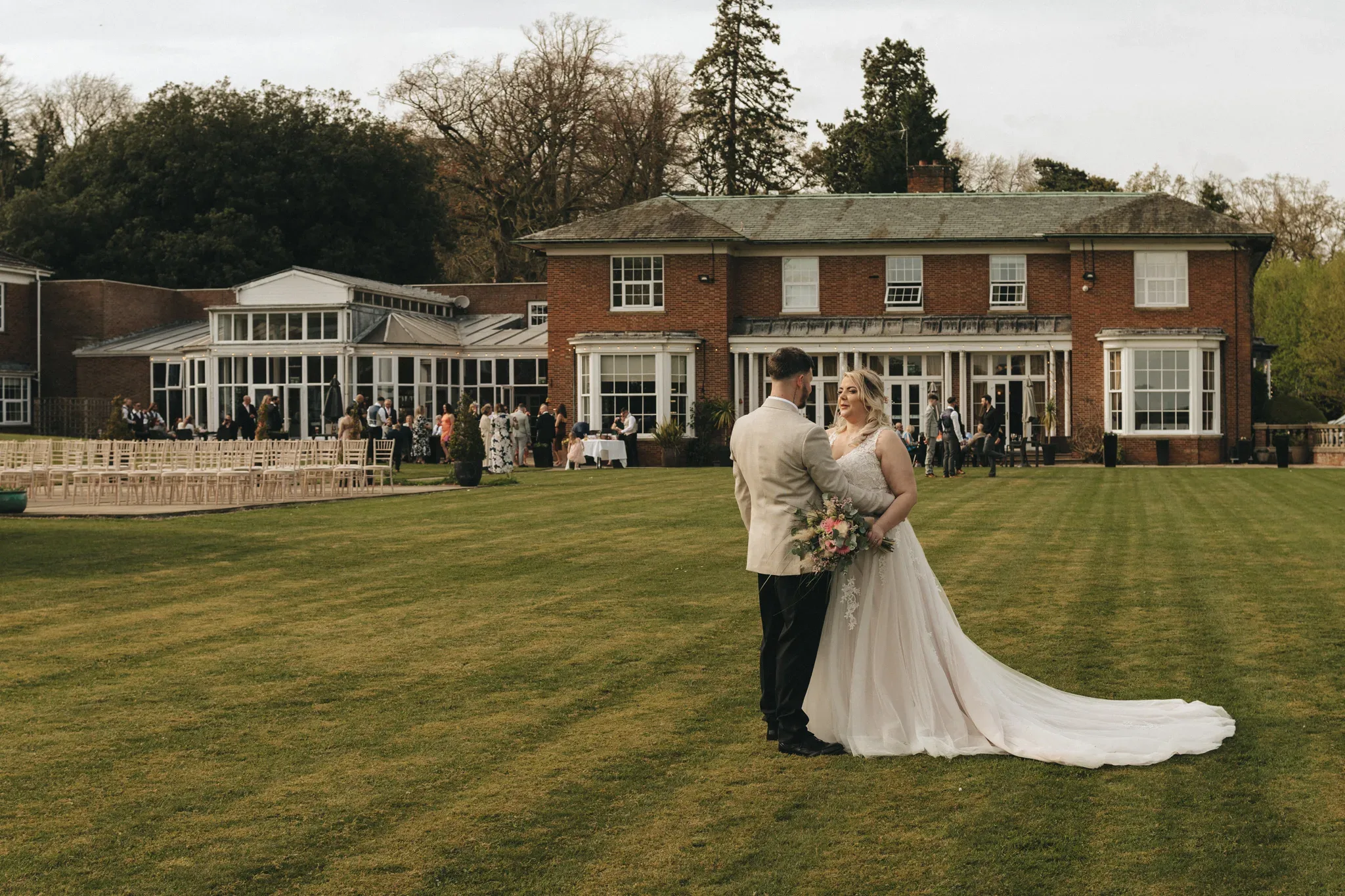 A bride in a white dress and a groom in a beige suit stand on a lush green lawn at Kenwick Park, Louth, holding hands and facing each other with a wedding party and a stately house in the background.