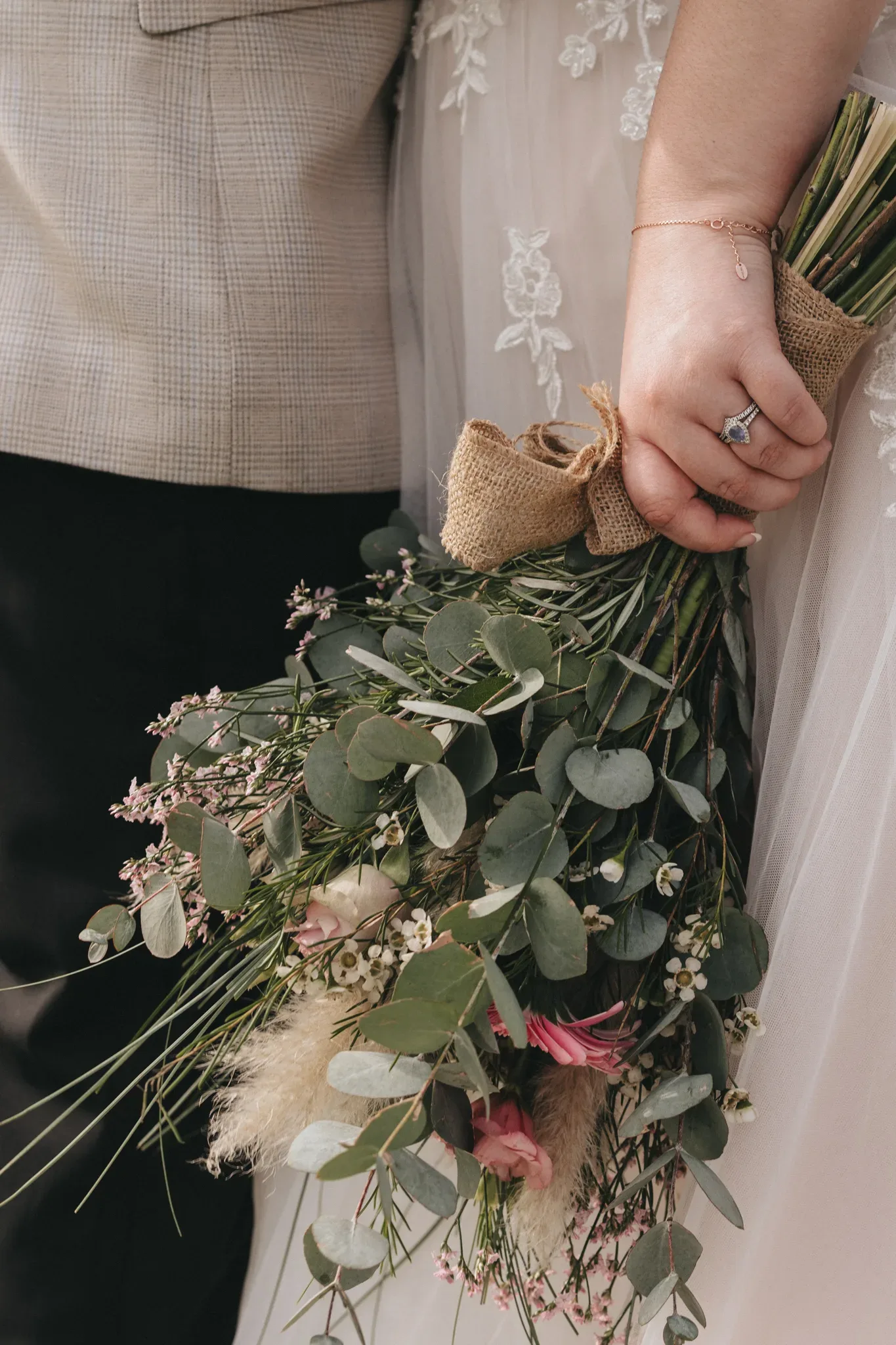 A close-up image of a bride and groom holding hands, the groom in a blazer and the bride in a lace gown. the bride holds a bouquet with eucalyptus leaves and pink flowers, showcasing a ring on her finger.