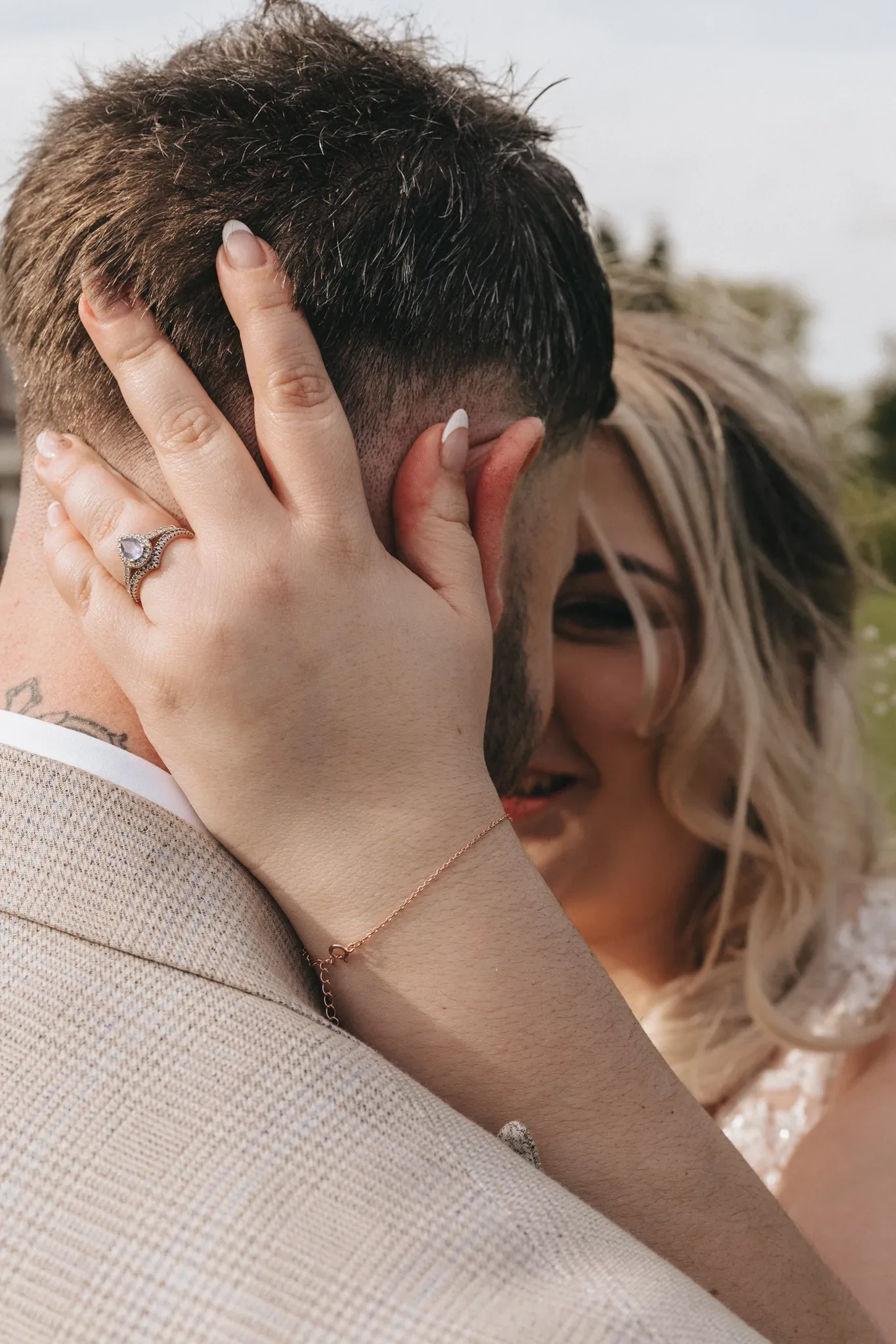 A bride, joyfully blurred in the background, holds the head of the groom, whose back is to the camera. the bride's hand, adorned with a diamond ring, gently touches his short, dark hair. warm, soft lighting enhances the intimate moment.