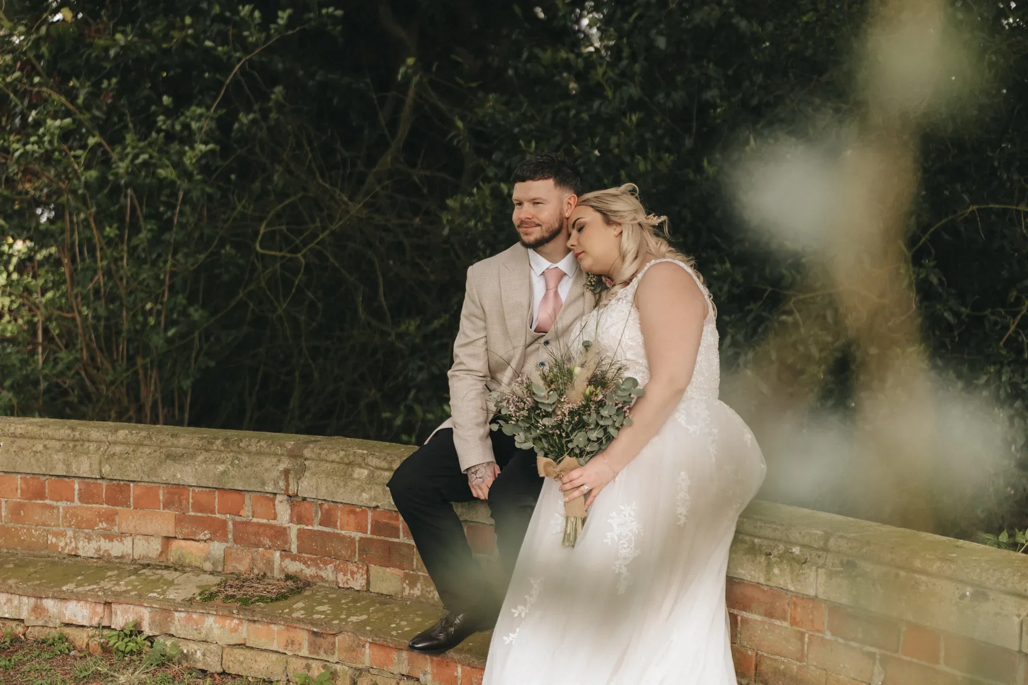 A bride and groom sit on a brick wall in a garden setting, the bride adjusting her dress, holding a bouquet, with the groom looking at her smiling. both wear formal wedding attire. green foliage surrounds them.