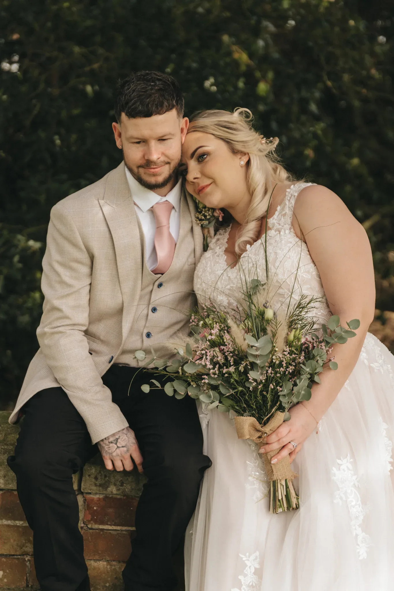 A bride and groom sit closely together, smiling softly, in a garden. the bride, on the right, holds a bouquet with green and purple flowers. both are dressed formally, the groom in a light beige suit with a pink tie, and the bride in a white lace gown.