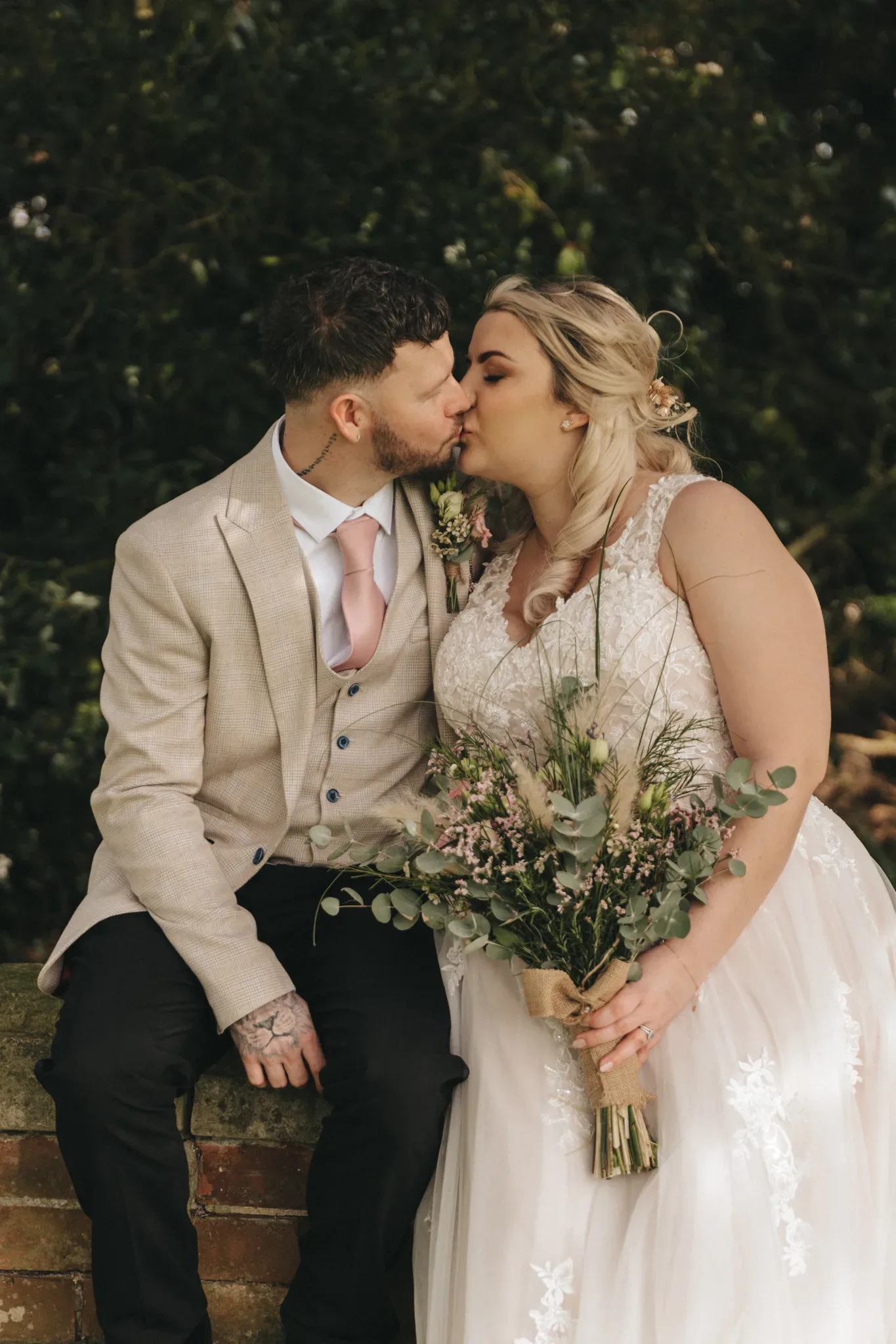 A bride and groom sharing a kiss while sitting on a brick wall. the bride holds a bouquet of green and white flowers, and both are dressed in elegant wedding attire, the groom in a beige suit and the bride in a lace gown.