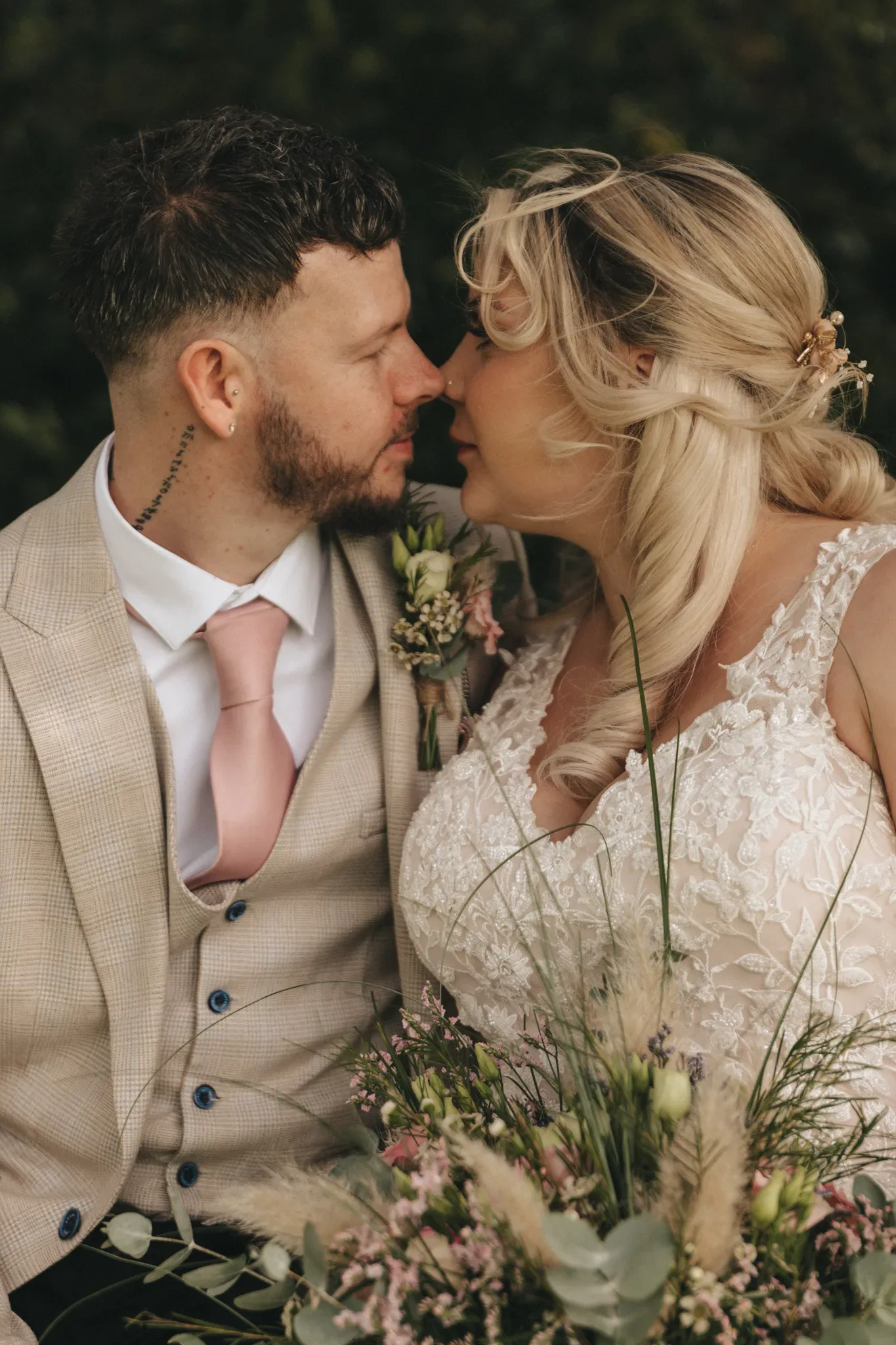 A bride and groom intimately touch foreheads, surrounded by greenery. the bride, in a lace wedding dress, has a delicate flower in her hair, while the groom wears a beige suit and pink tie.