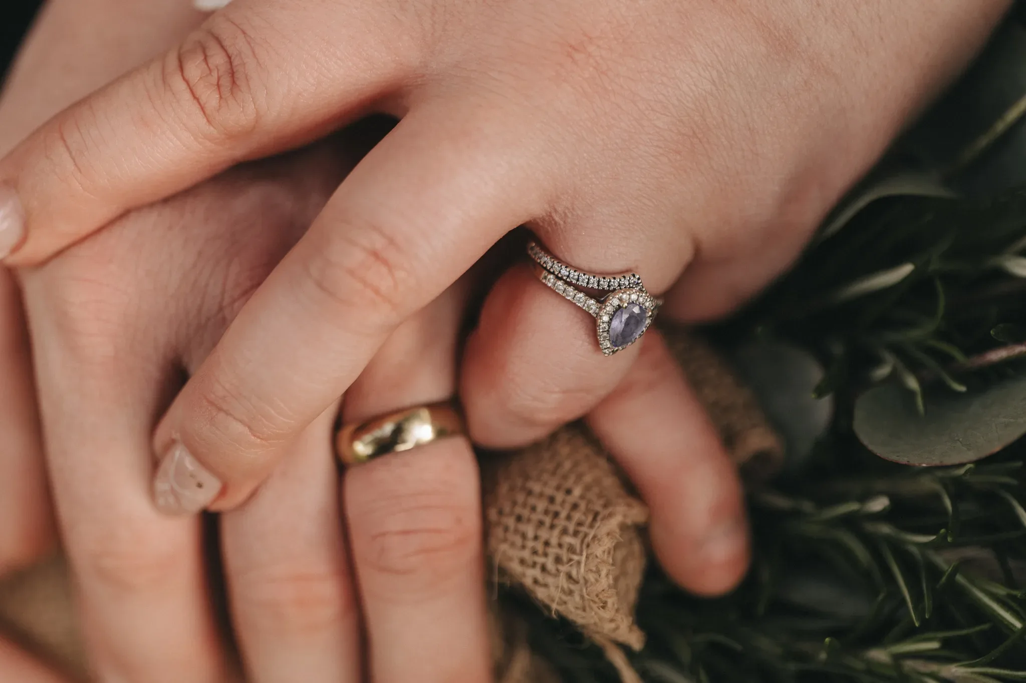 Close-up of a couple's hands gently touching, showcasing their wedding rings. the woman's ring has a prominent purple gemstone surrounded by smaller diamonds, while the man's is a simple gold band.