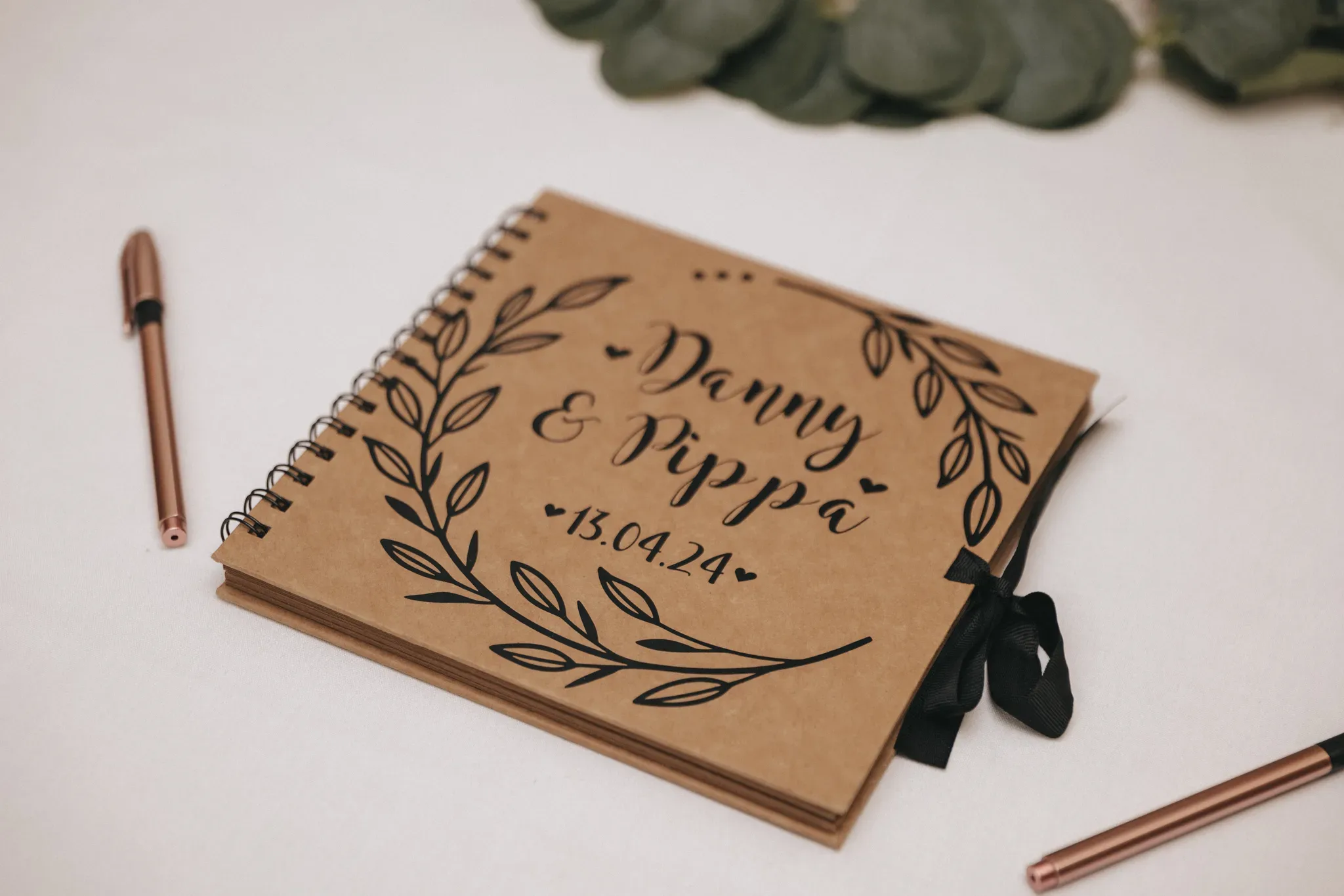 A personalized notebook with a brown cover, titled "danny & myra * 13.09.14" in decorative script, surrounded by a floral design, lies on a white surface next to a copper pen and green leaves.