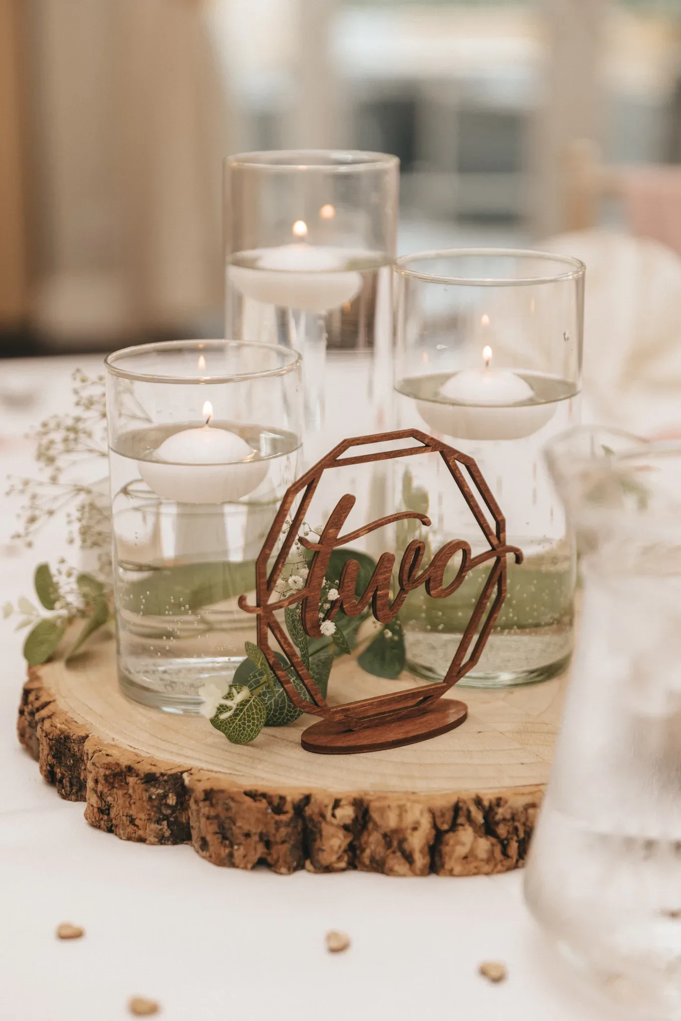 A rustic wedding table centerpiece featuring three glass votive candle holders with lit candles, set on a circular wooden slice. a decorative metal piece with the word "love" in script is centered among the candles, surrounded by small green leaves.