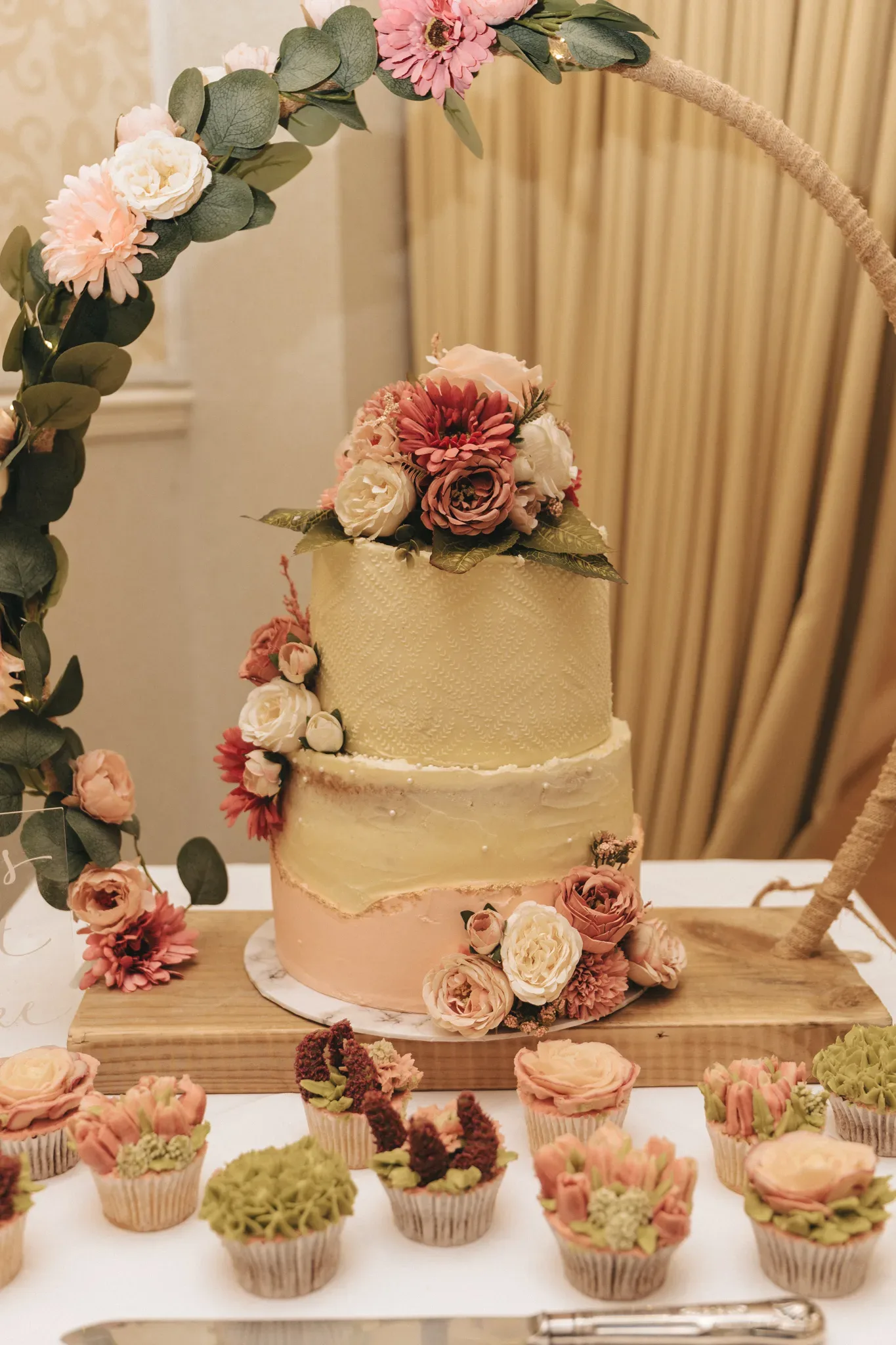 A three-tiered wedding cake adorned with pink and cream flowers, displayed on a table surrounded by matching floral cupcakes, set against a soft, beige backdrop.