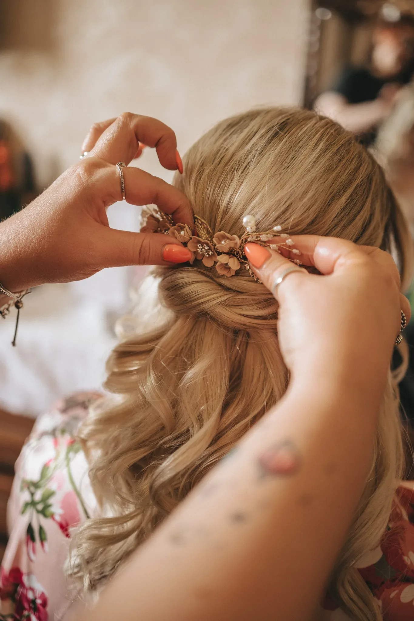 A person fastens a decorative floral hair accessory into another person's intricately styled blonde hair, featuring curls and braids. the setting appears to be a room with a soft-focus background.