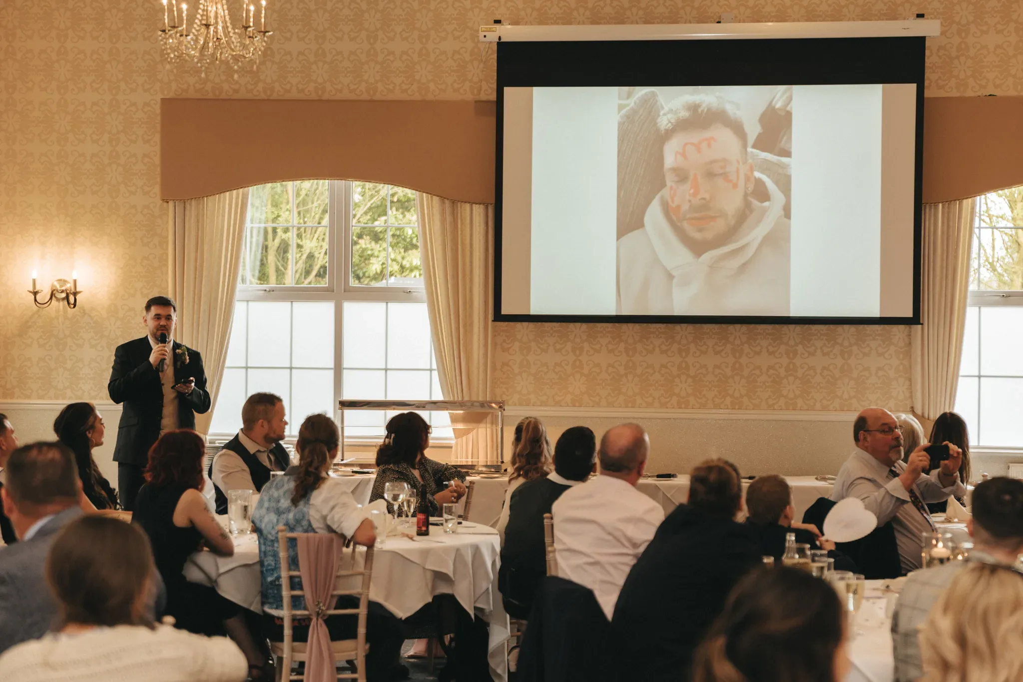 An elegant room with guests seated at tables listening to a man giving a speech. behind the speaker, a large screen displays a humorous photo of a man with playful writing on his forehead. chandeliers hang from the ceiling, adding a sophisticated touch to the setting.