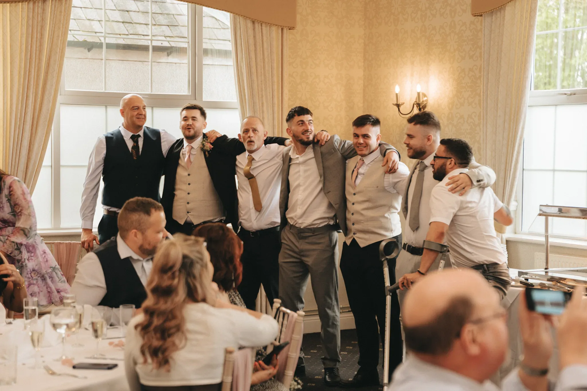 A group of six men embracing and posing happily together at a wedding reception. they are dressed in formal attire, with the room filled with seated guests. the decor includes elegant cream wallpaper and chandeliers.