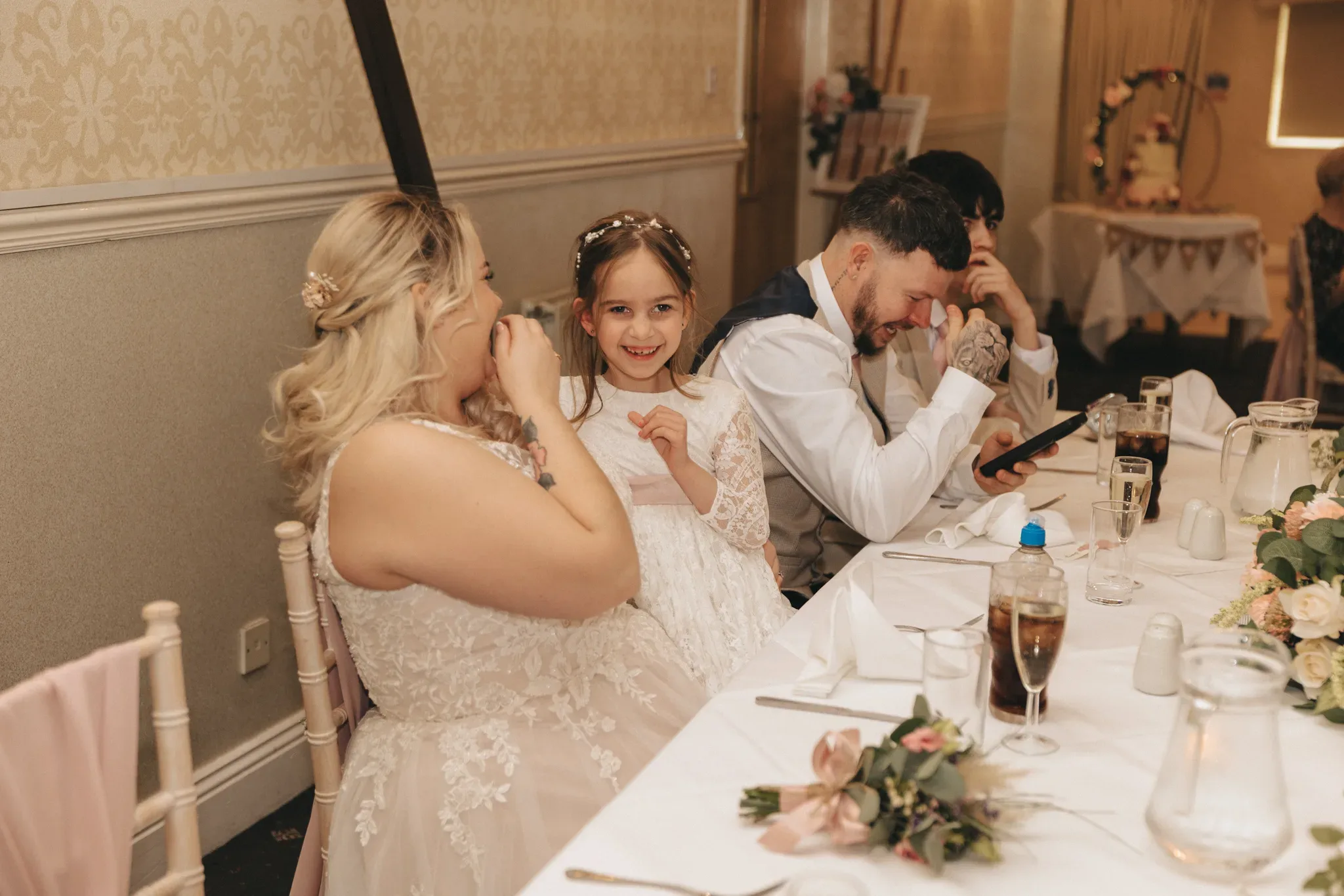 A joyful wedding reception scene with a young girl smiling broadly between two adults, a bride and a man likely checking his phone. they're seated at a table adorned with floral arrangements.