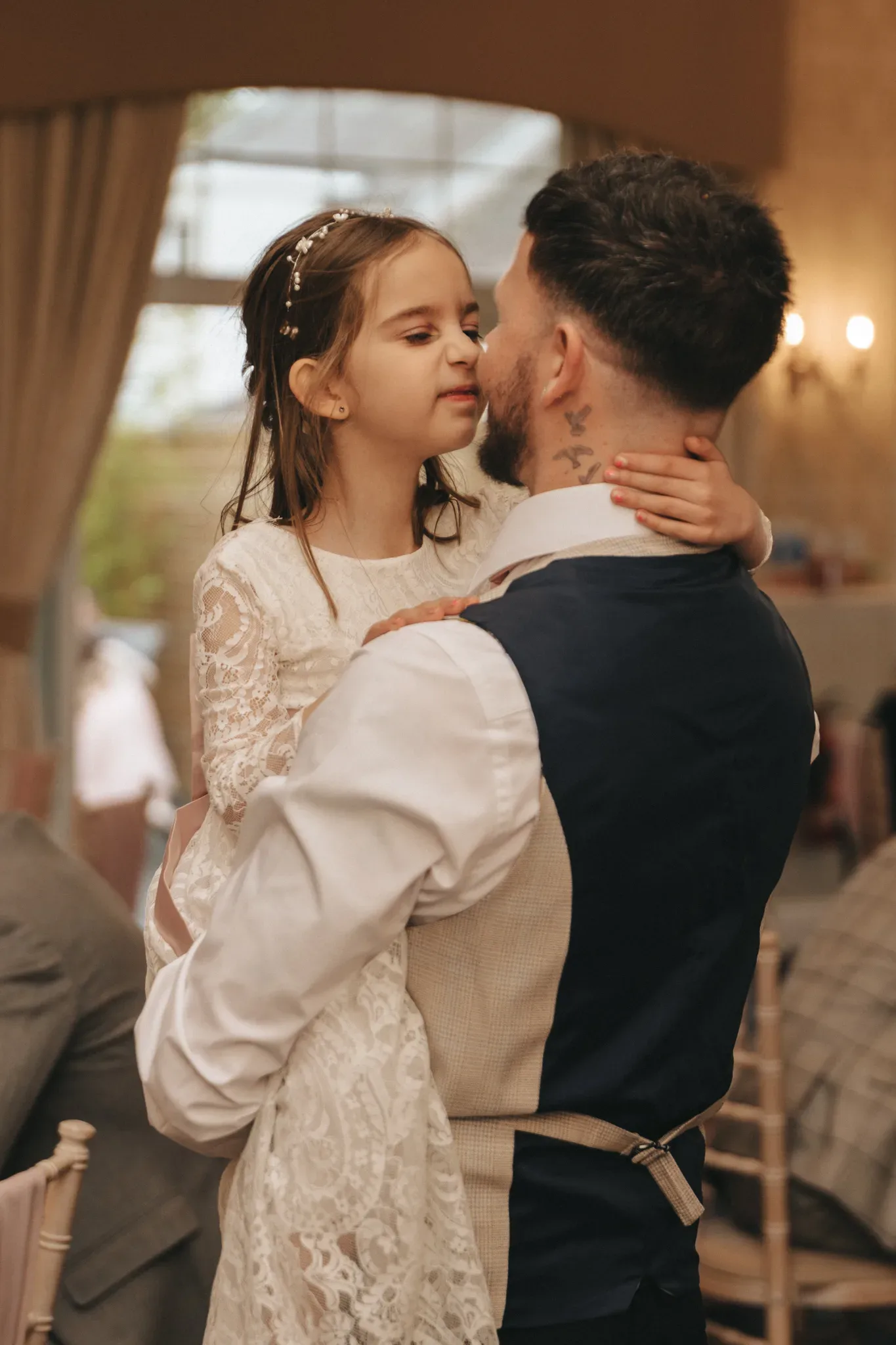A young girl in a white lace dress embraces a bearded man in a formal vest and rolled-up sleeves. she touches his cheek affectionately as they share a tender moment in a warmly lit indoor setting.