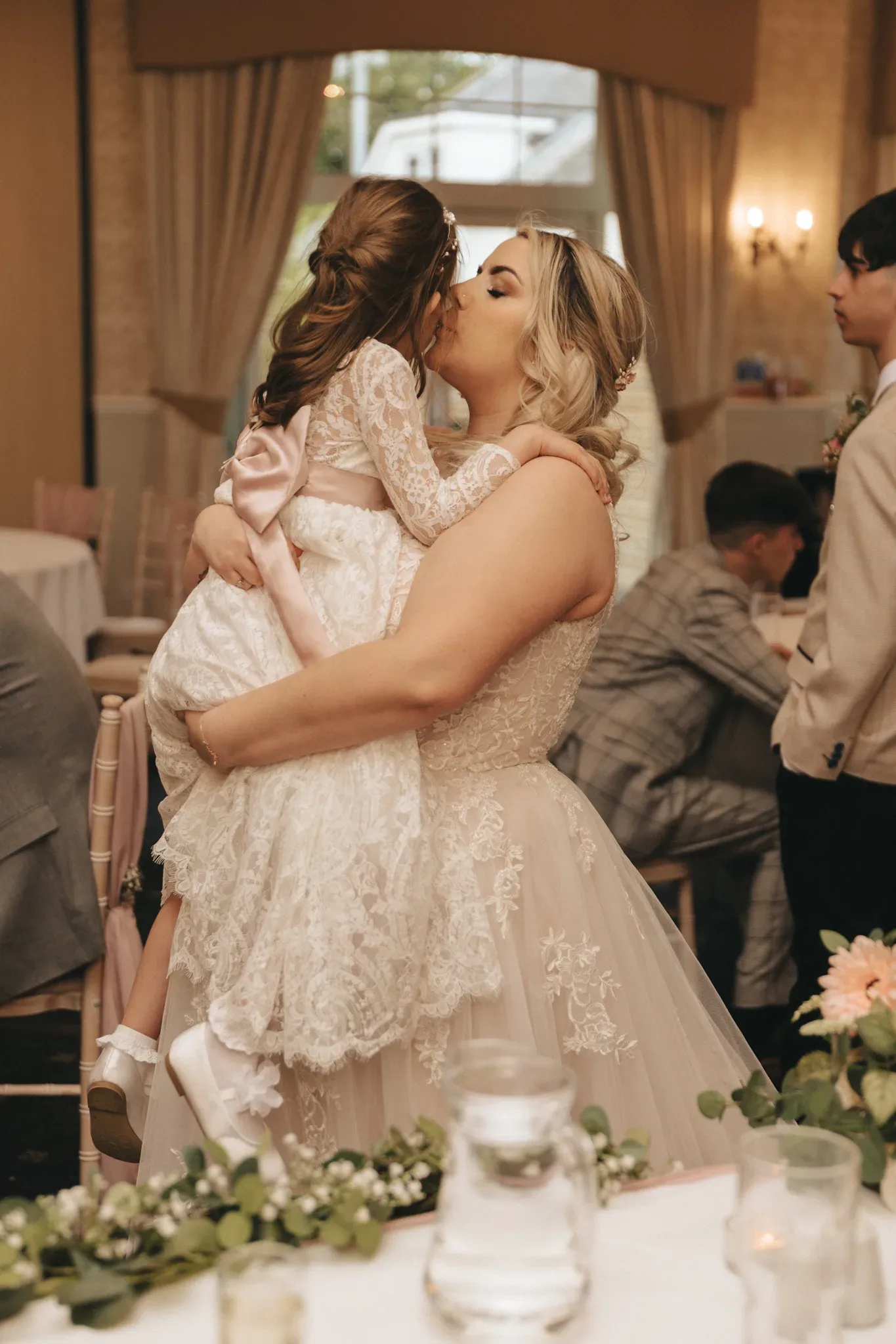 A bride in a lace gown lovingly lifts and kisses a young girl in a white lace dress at a wedding reception, surrounded by floral decorations and guests.