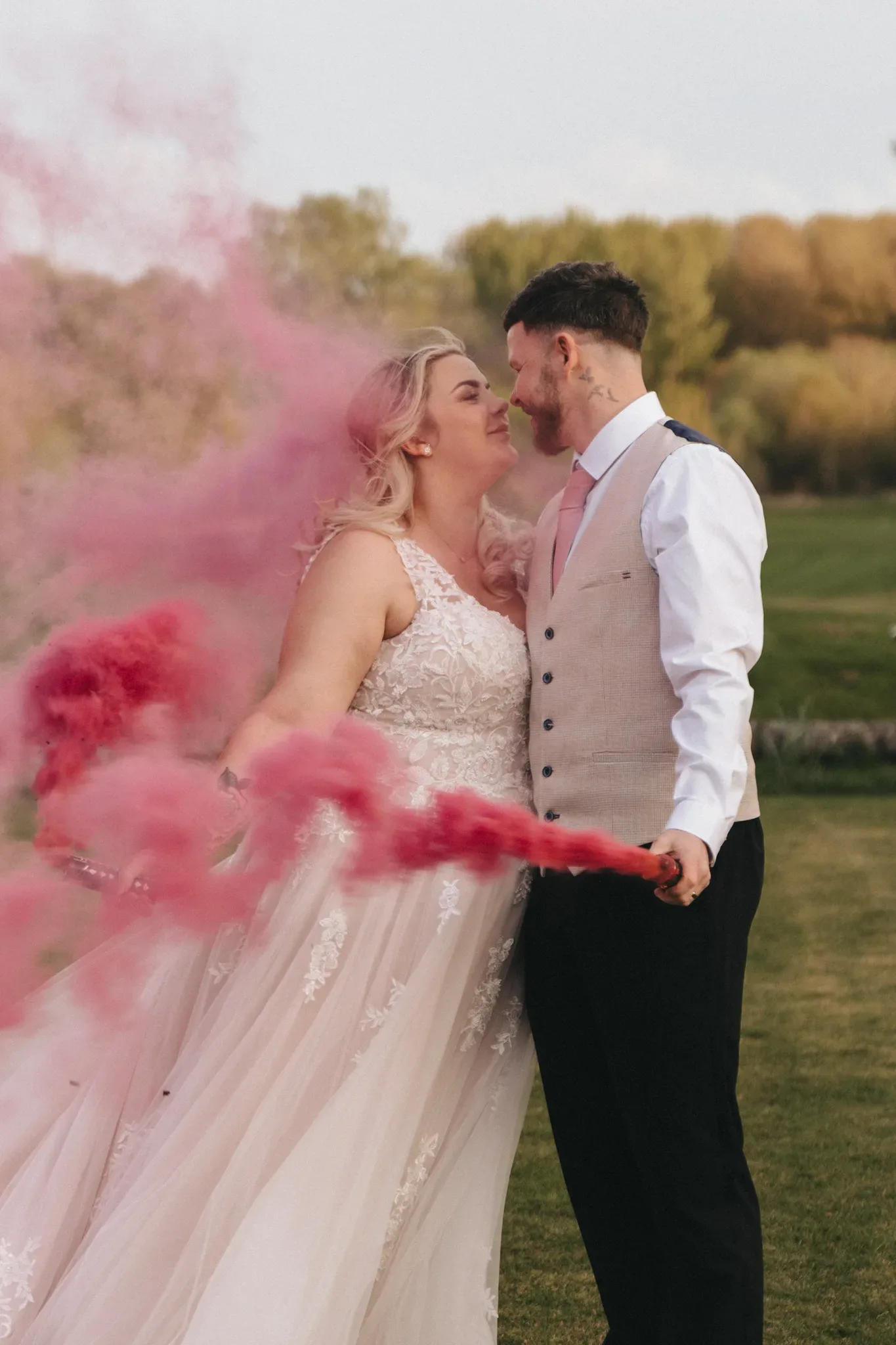 A bride and groom embrace outdoors with pink smoke swirling around them. the bride wears a white gown and the groom is in a gray vest and white shirt. they gaze affectionately at each other with a grassy field and trees in the background.