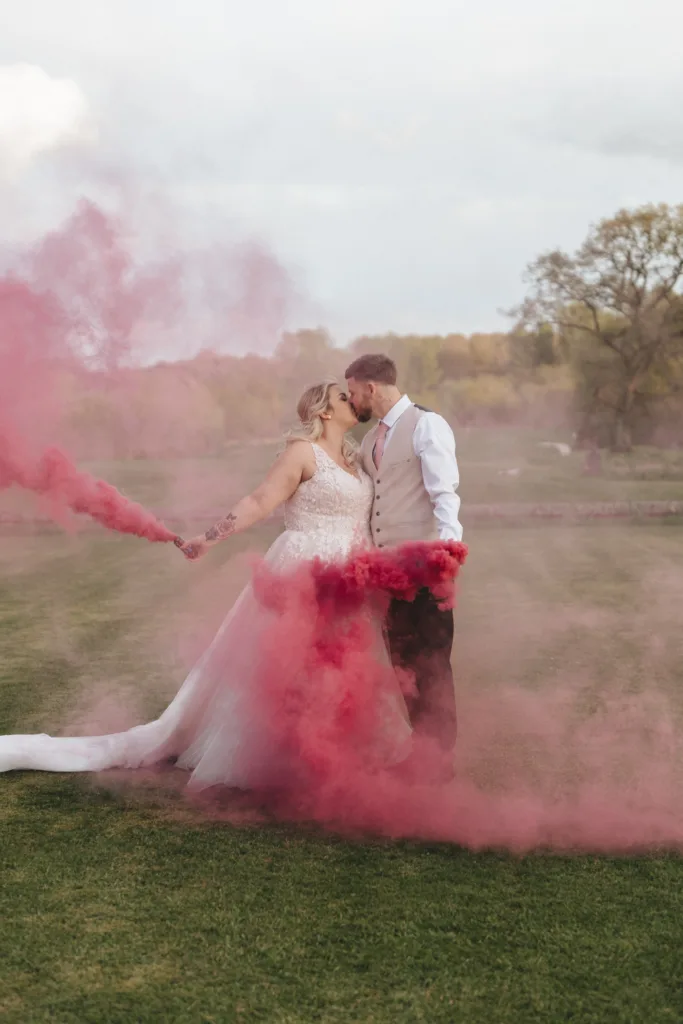 A bride and groom share a kiss surrounded by pink smoke on a grassy field. the bride wears a white gown and holds a smoke bomb; the groom is in a beige suit. the setting is serene with trees in the background.