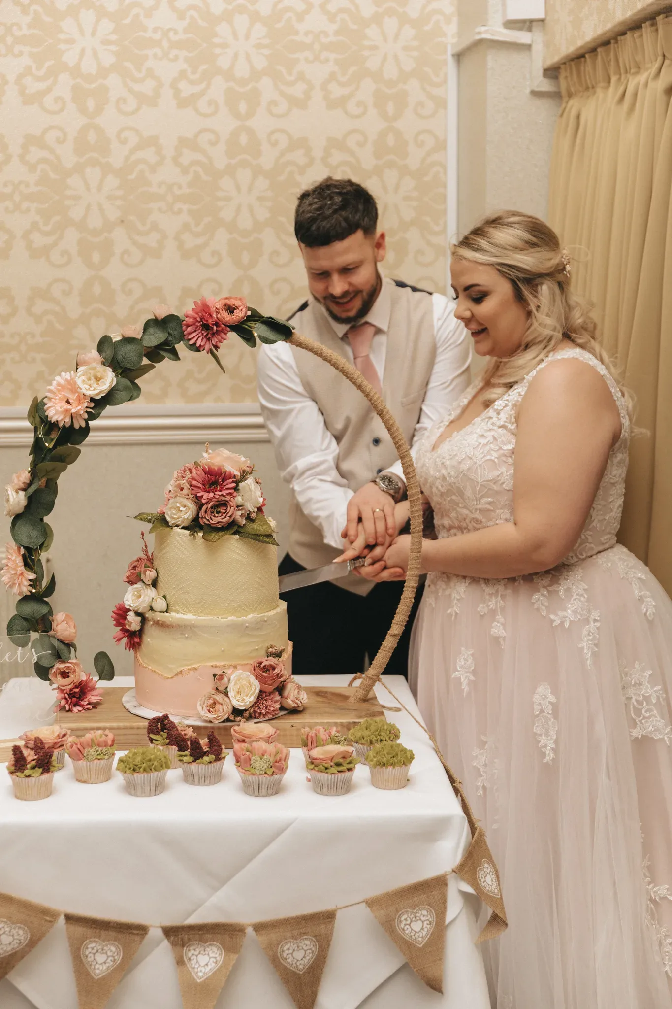 A bride and groom are cutting a three-tier wedding cake adorned with pink flowers, surrounded by cupcakes, under a floral arch, inside a warmly lit room with elegant wallpaper.