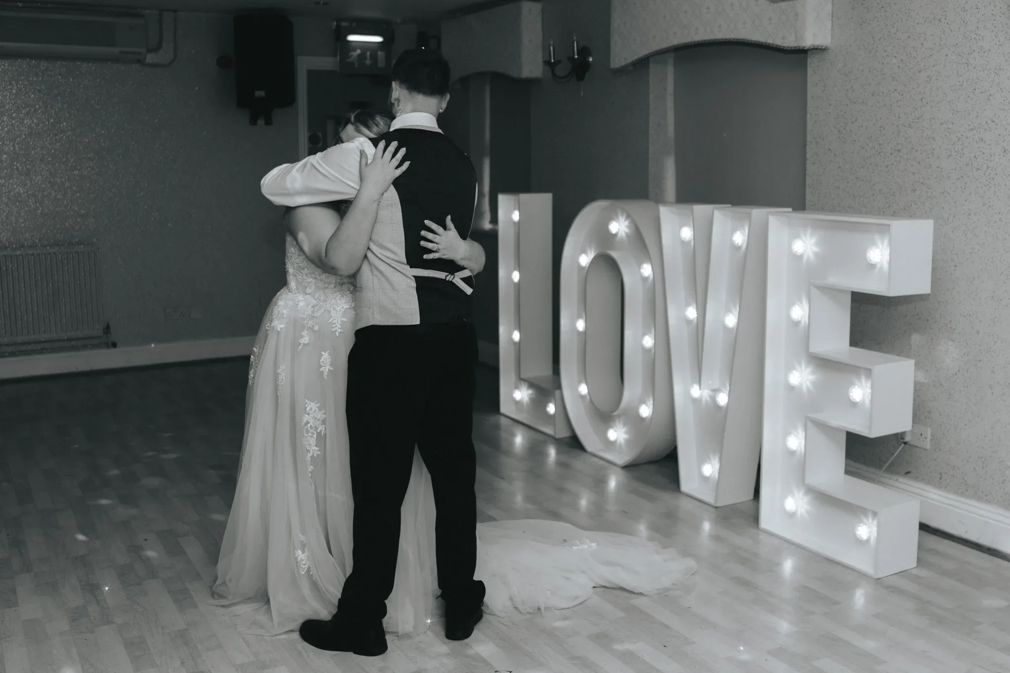 A couple shares a slow dance at their wedding reception, surrounded by large illuminated letters spelling "love." the bride, in a long gown with a train, embraces the groom from behind. the venue has a simple, elegant decor.