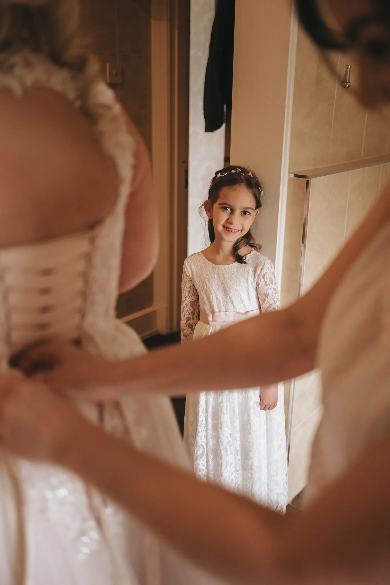 A young girl in a white lace dress smiles as she looks at a bride being helped into her gown, visible in a mirror's reflection. the setting suggests a room prepared for a wedding.