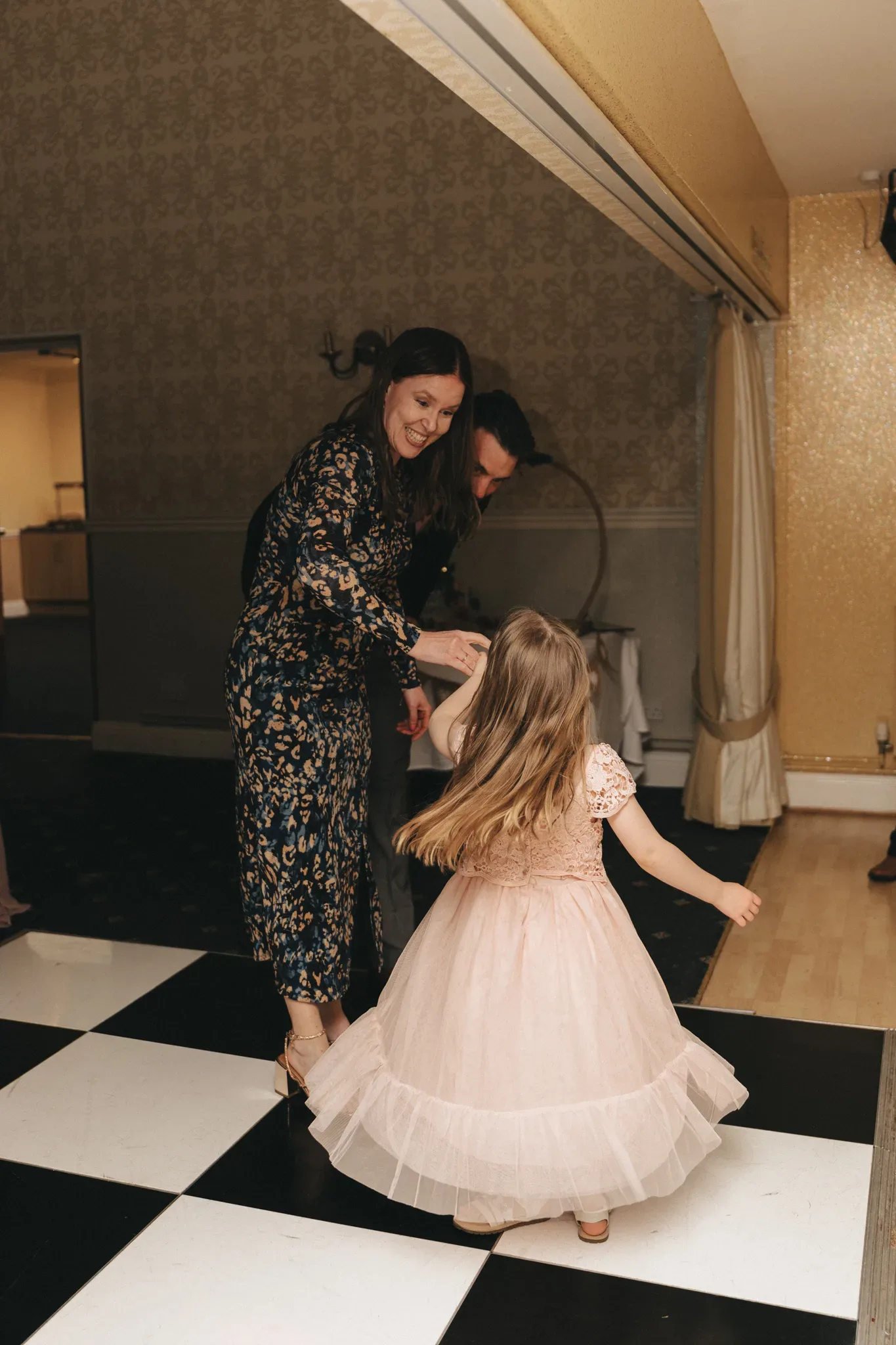 A woman in a floral dress and a man playfully dance with a young girl in a pink tutu on a black and white checkered floor, in a warmly lit room with vintage wallpaper. they are all smiling, creating a joyful, festive atmosphere.