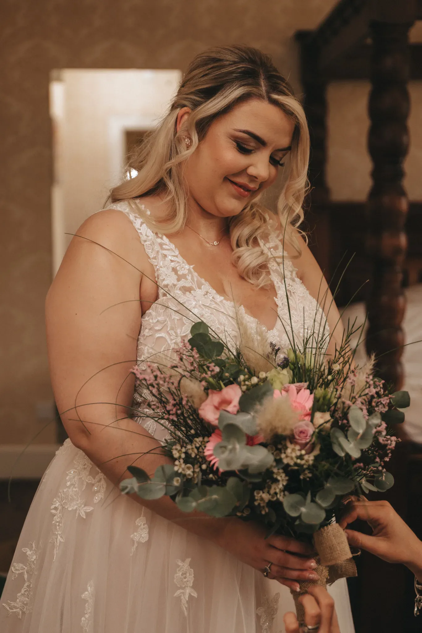 A radiant bride in a lace wedding dress gazes affectionately at her bouquet of pink, white, and green flowers, with warm lighting enhancing the elegant indoor setting.